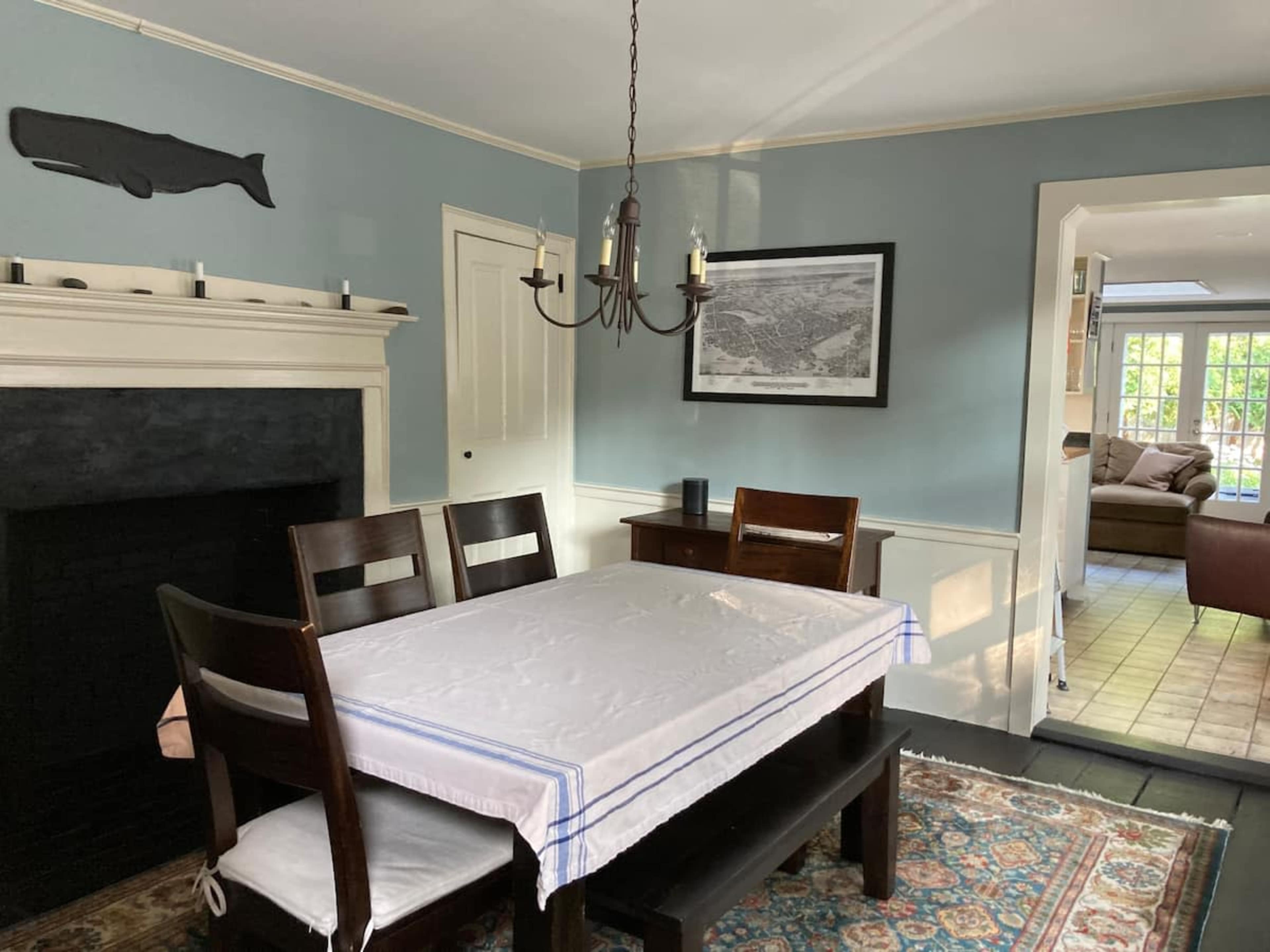 A dining room features a wooden table with four chairs, a chandelier above, and a light blue wall with a whale decoration.