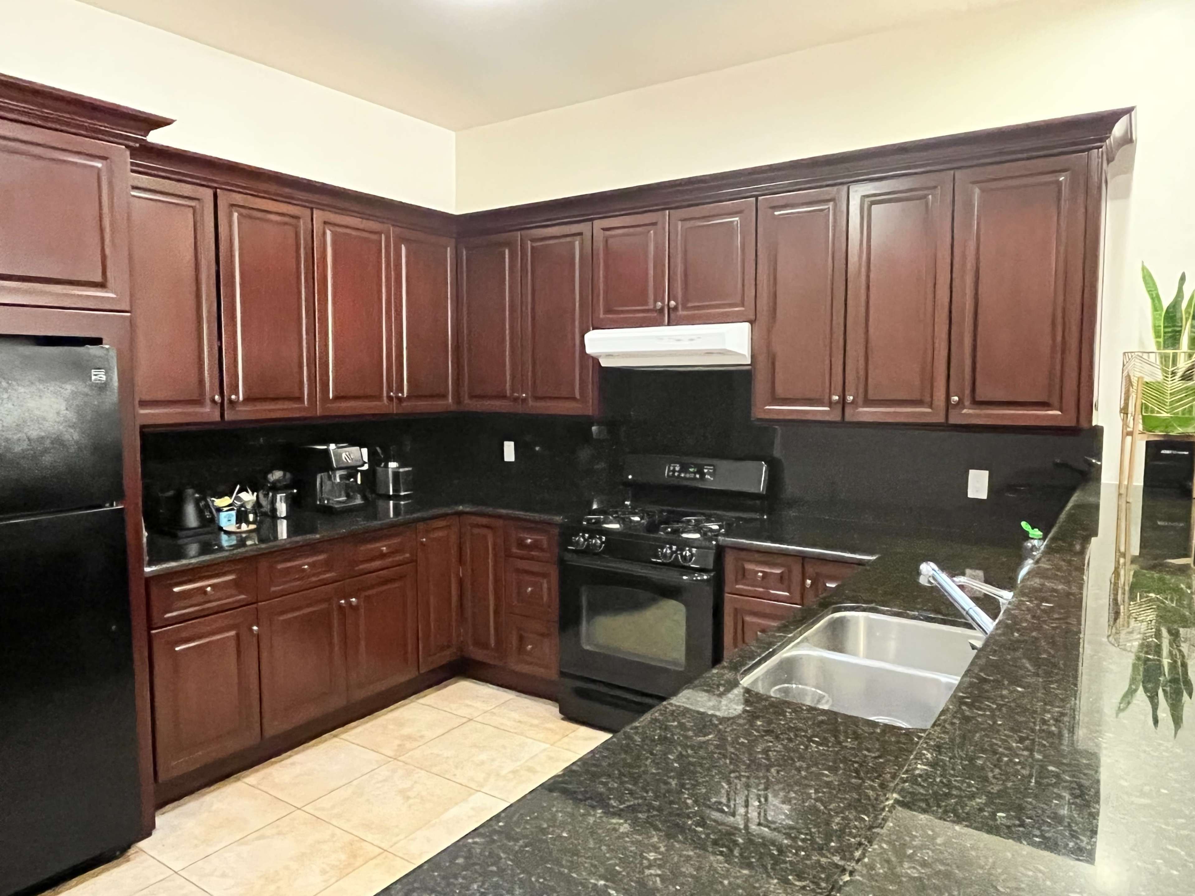 The image shows a spacious kitchen featuring dark wood cabinetry, a black refrigerator, a black stove, and a granite countertop with a sink.