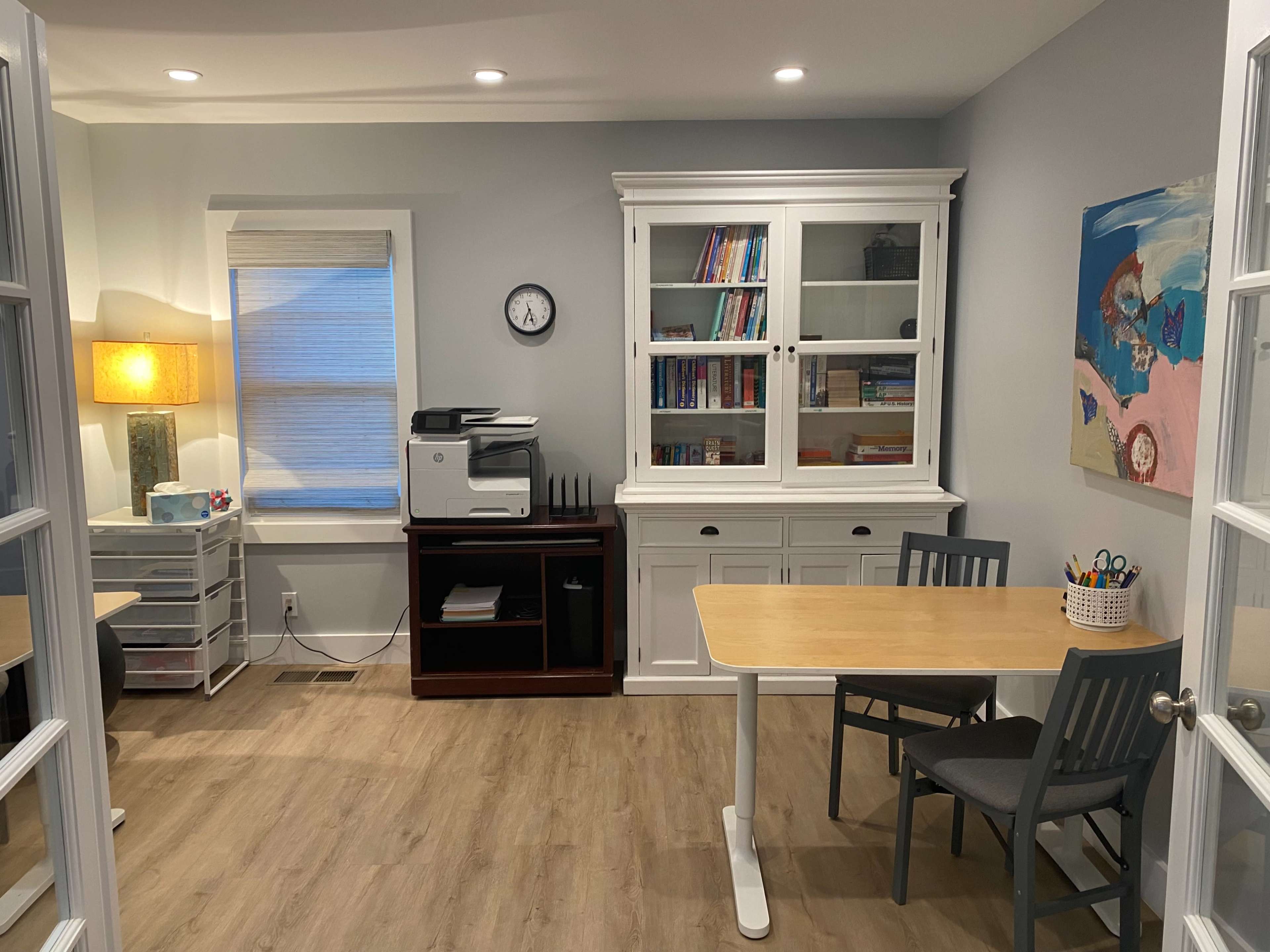 A modern, well-lit office space featuring a printer, a white bookshelf filled with books, a table with chairs, and a colorful artwork on the wall.