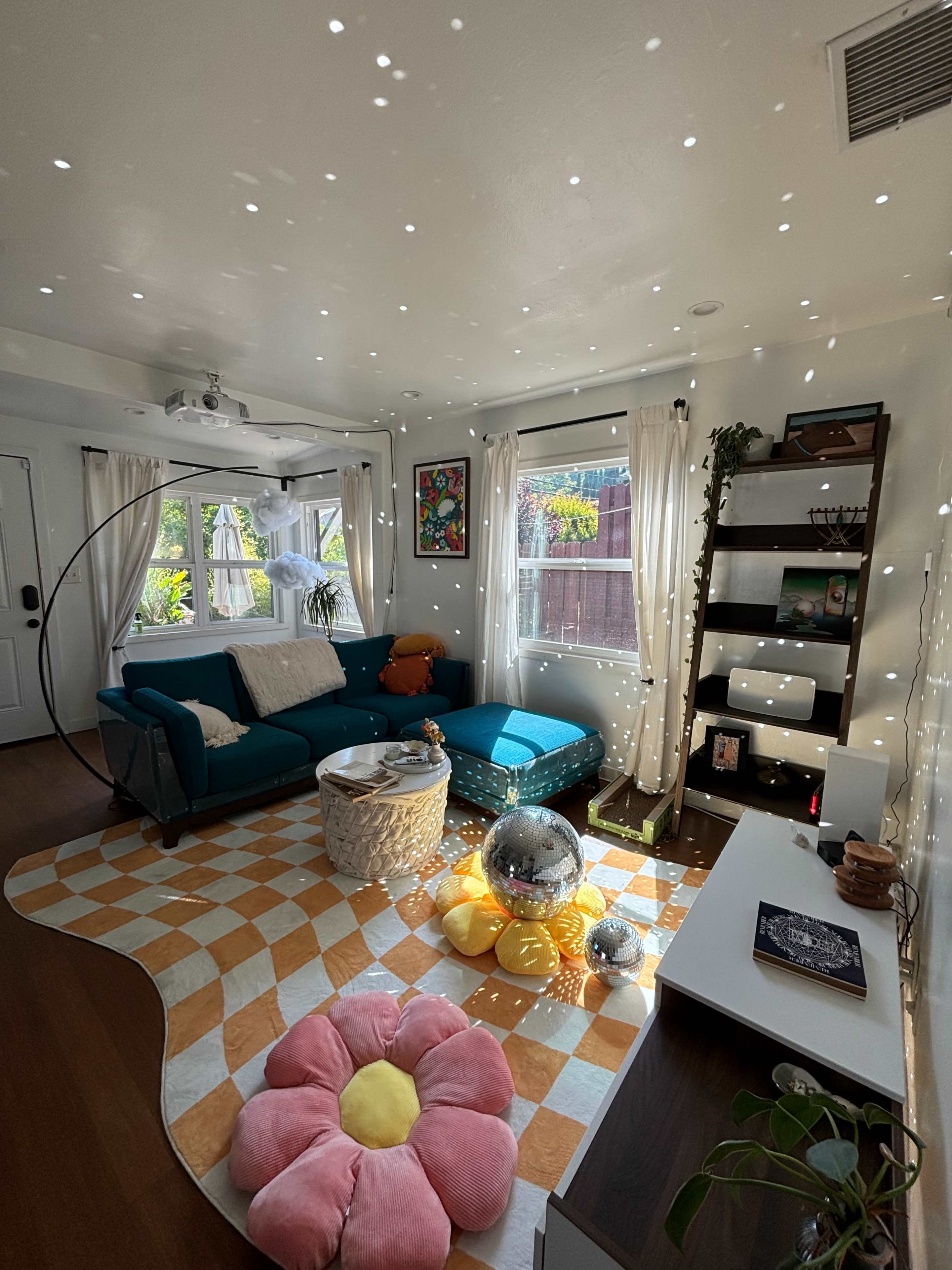 A brightly lit living room features a teal couch, a floral rug, and a disco ball reflecting light across the walls.