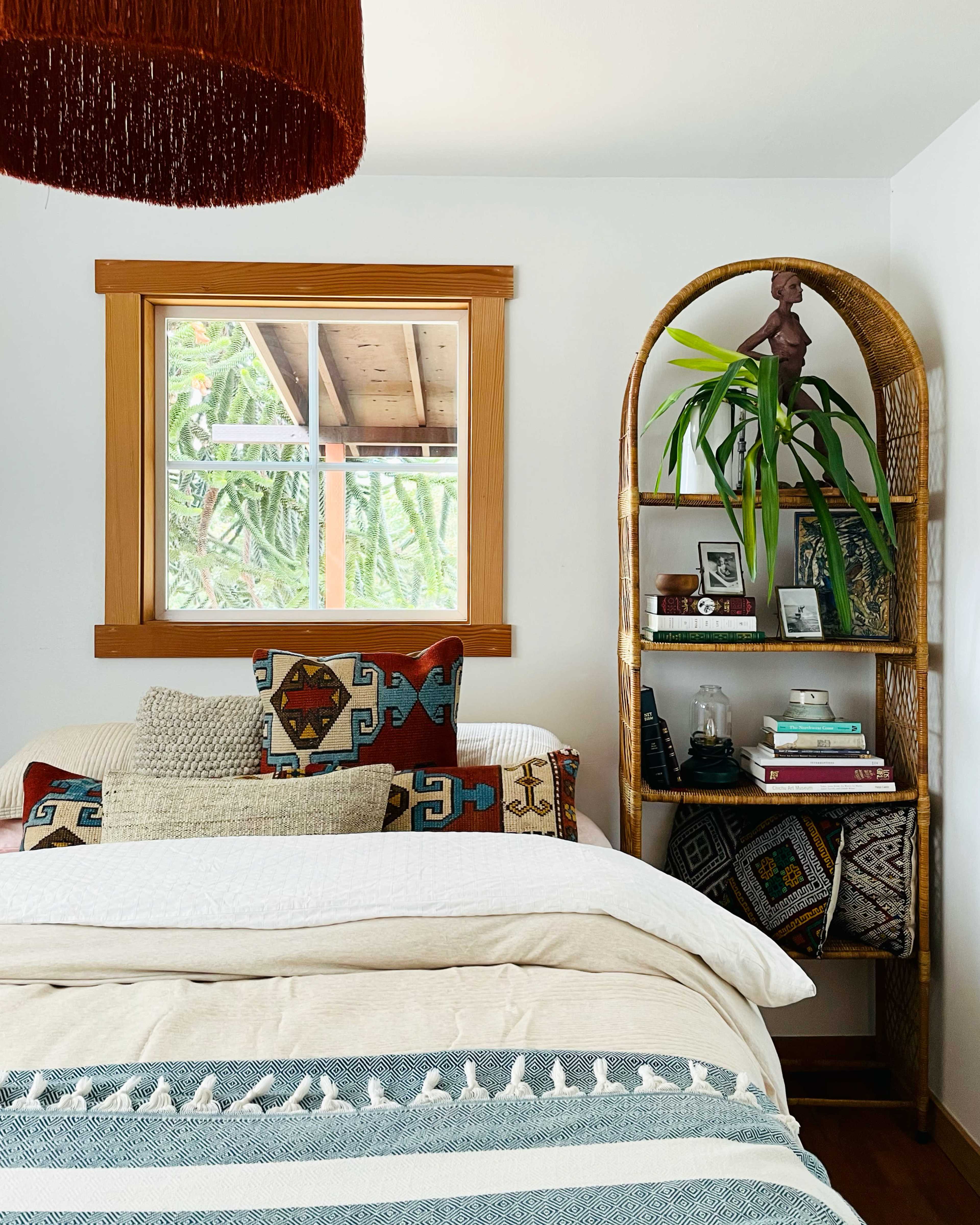 A cozy bedroom with a bed adorned with various patterned pillows, a shelf filled with books and decor, and a window allowing natural light to enter.