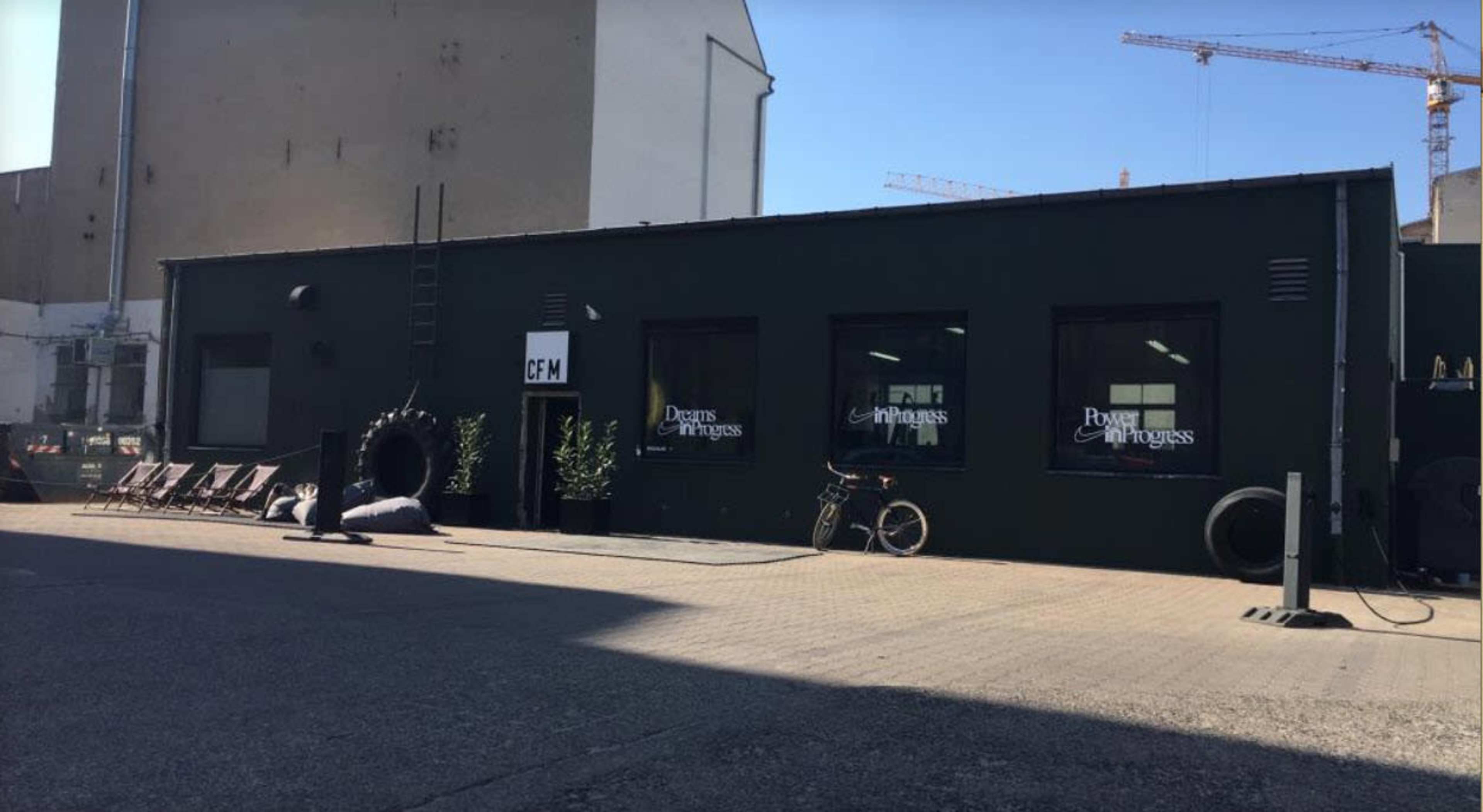 A green building with large windows and a flat roof, situated in an industrial area alongside several bicycles and outdoor furniture.