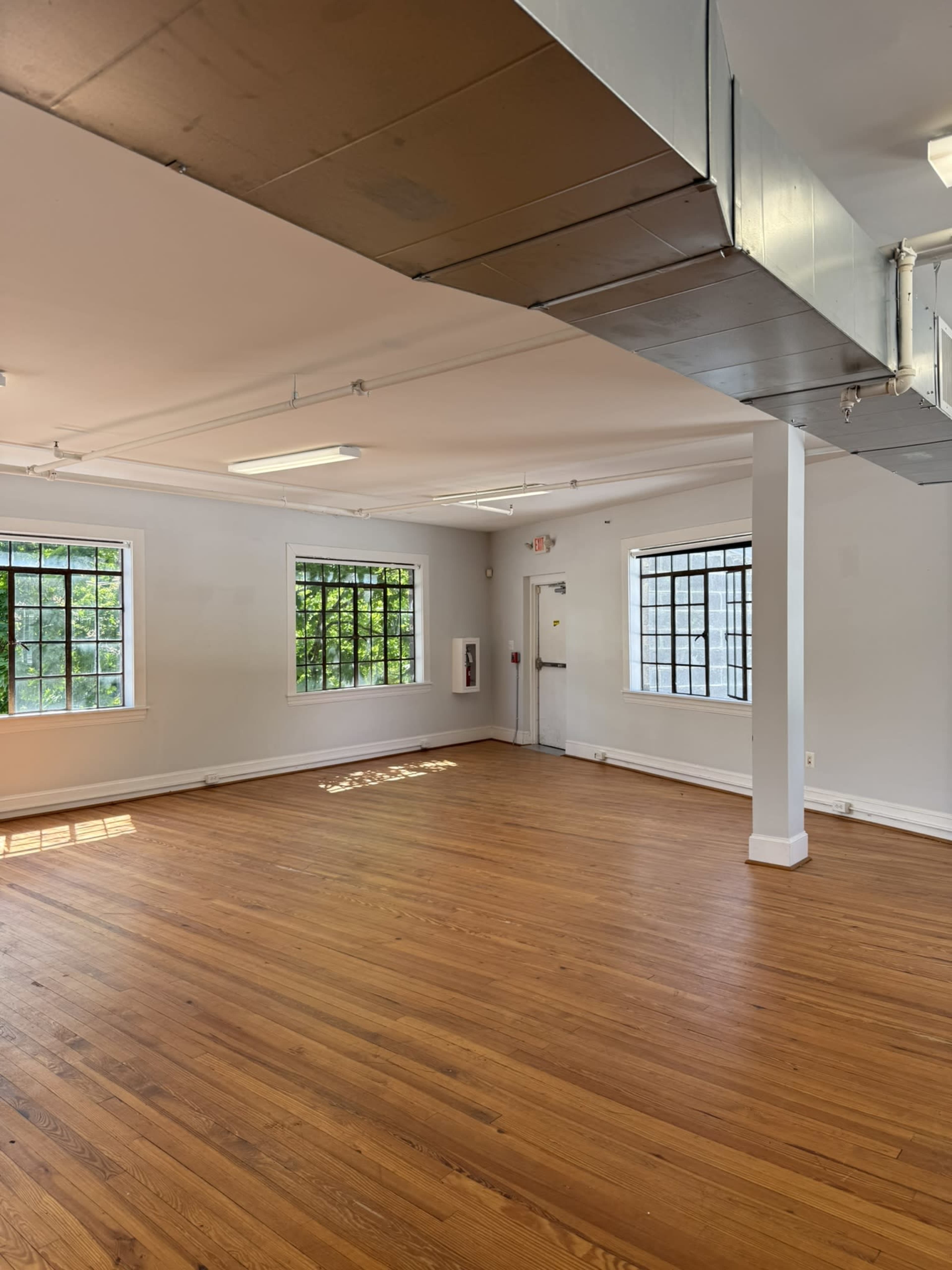 The image shows an empty room with polished wooden floors, large windows, and a door, featuring white walls and exposed ductwork.