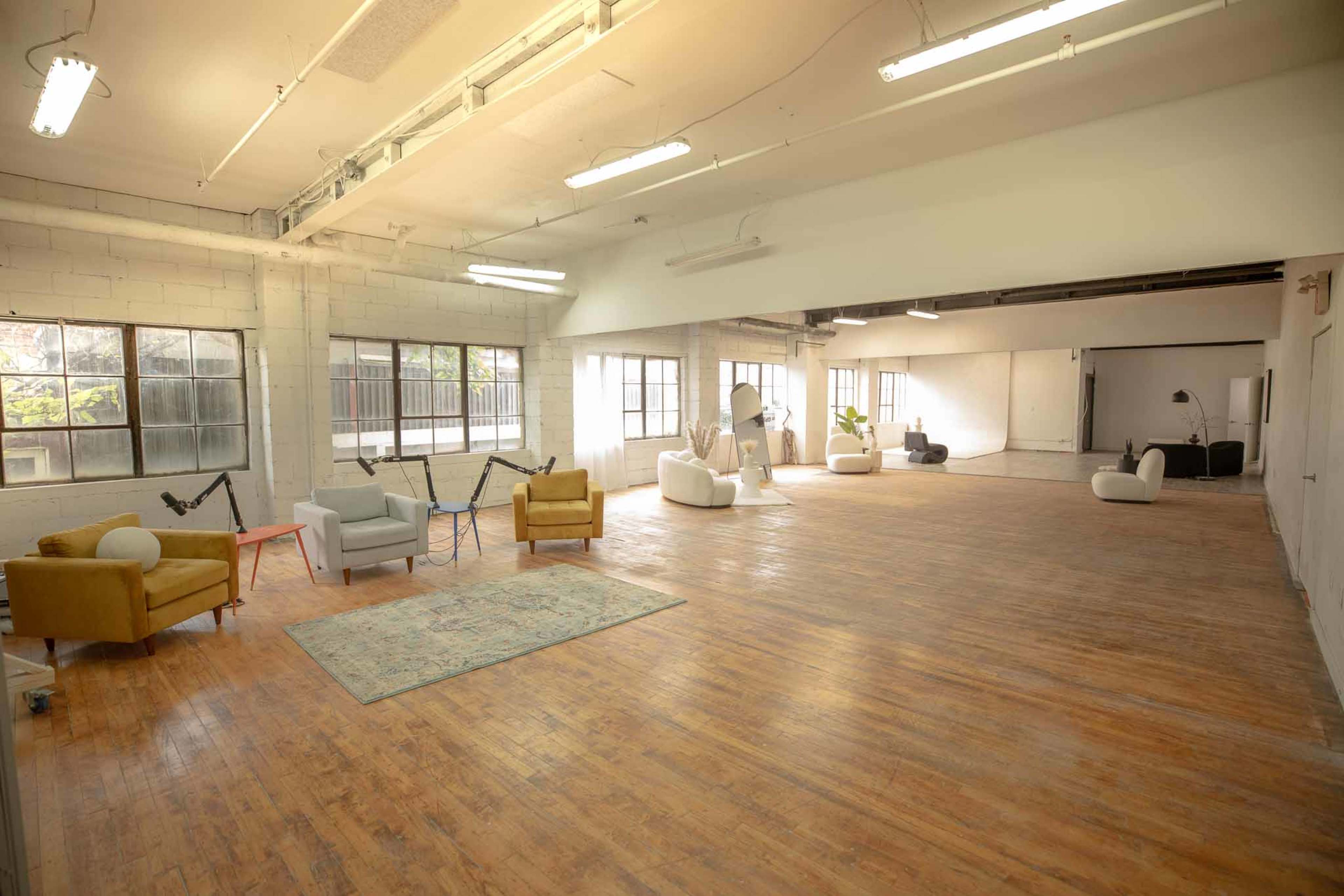 The image shows a spacious, well-lit room with wooden flooring, featuring various seating arrangements and large windows.