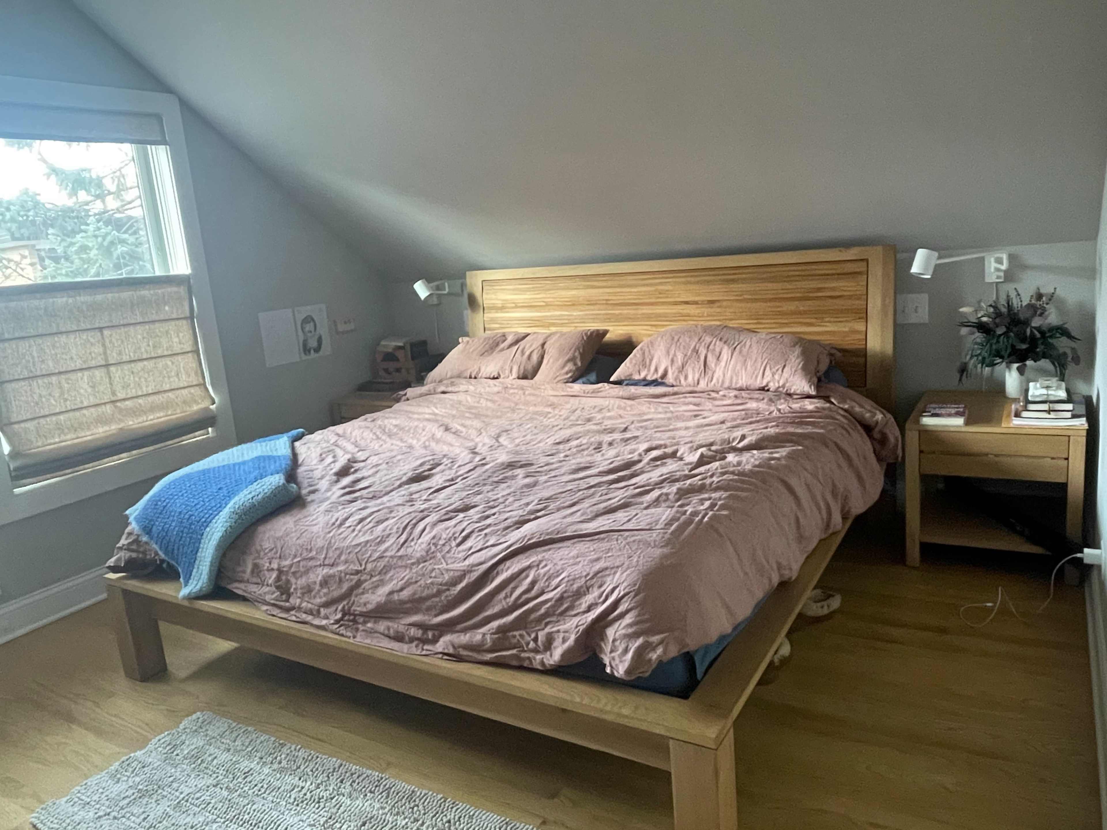 A cozy bedroom with a wooden bed, light brown bedding, a bedside table with books, and a window with a woven shade.