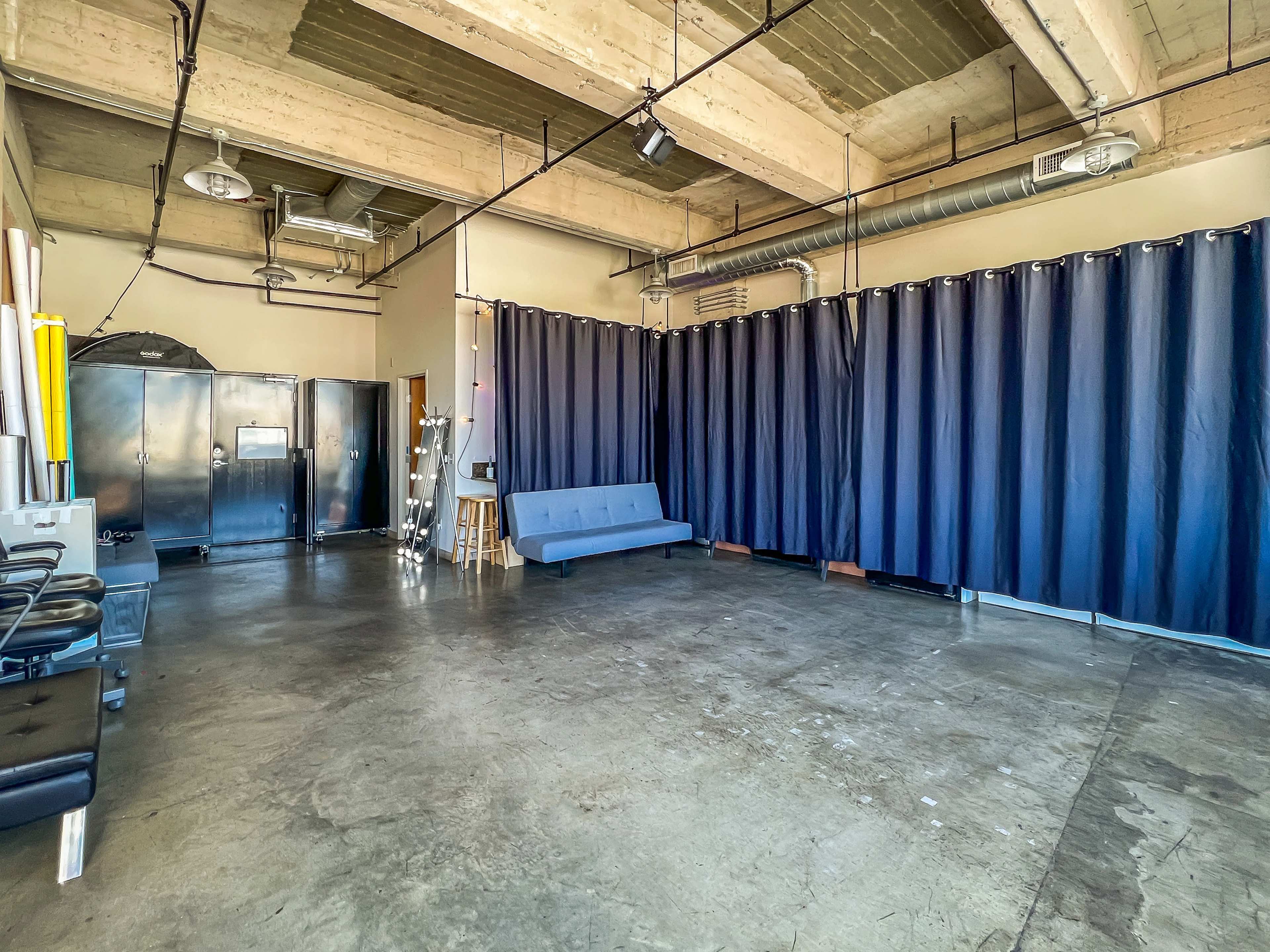 A spacious room with exposed ceilings, a concrete floor, a blue curtain on one side, a gray sofa, and cabinets along the back wall.