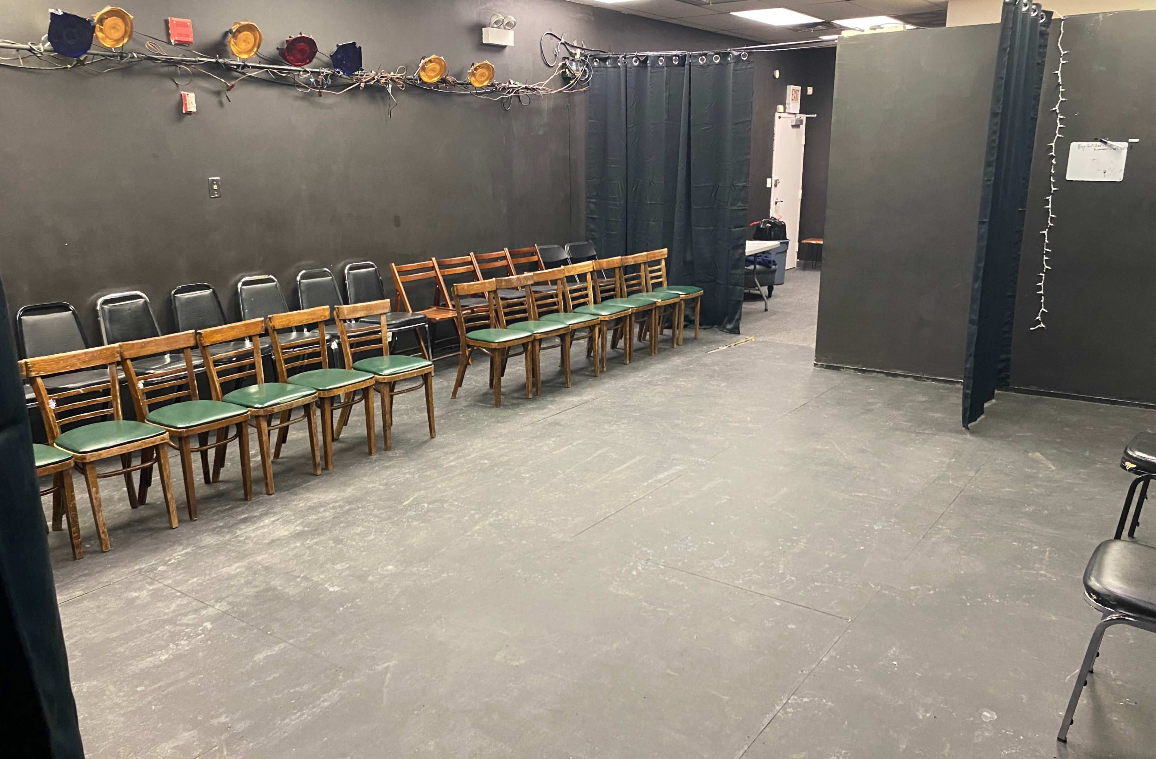 The image shows a room with a row of wooden chairs arranged facing a plain, dark wall, and partitions creating separate spaces.