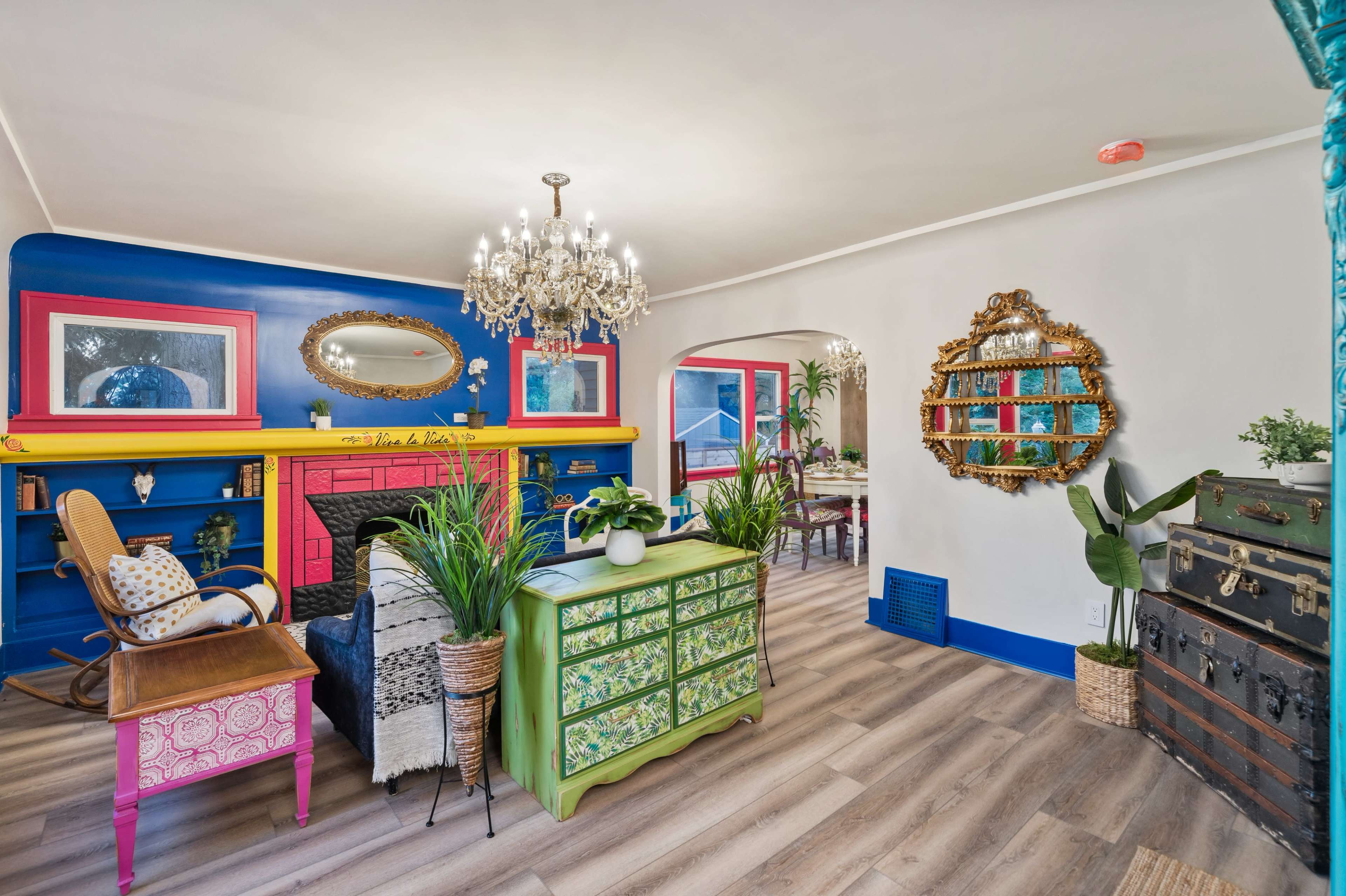 A colorful living room features a vibrant blue and pink fireplace, a chandelier, decorative plants, and eclectic furniture.