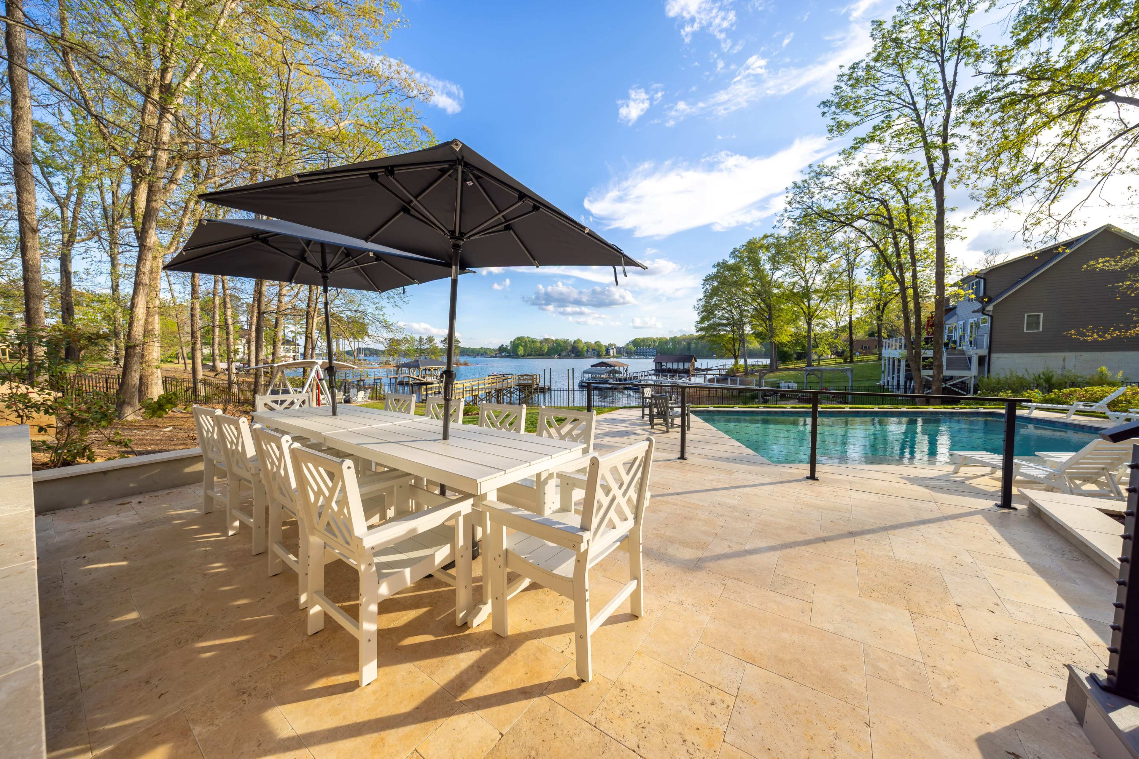 The image shows a patio area with a large white dining table and chairs under a black umbrella, overlooking a pool and a lake surrounded by trees.