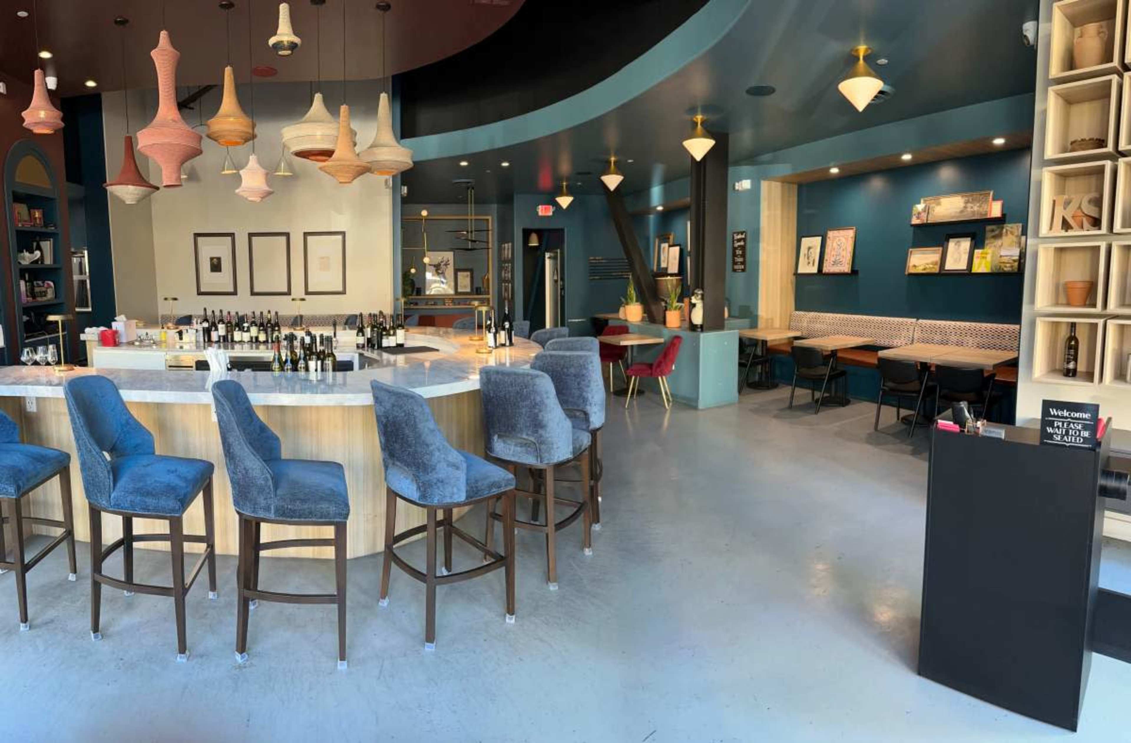 The image shows a modern bar interior with a curved counter, high stools, and a mix of seating options around the space, featuring colorful pendant lights and artwork on the walls.