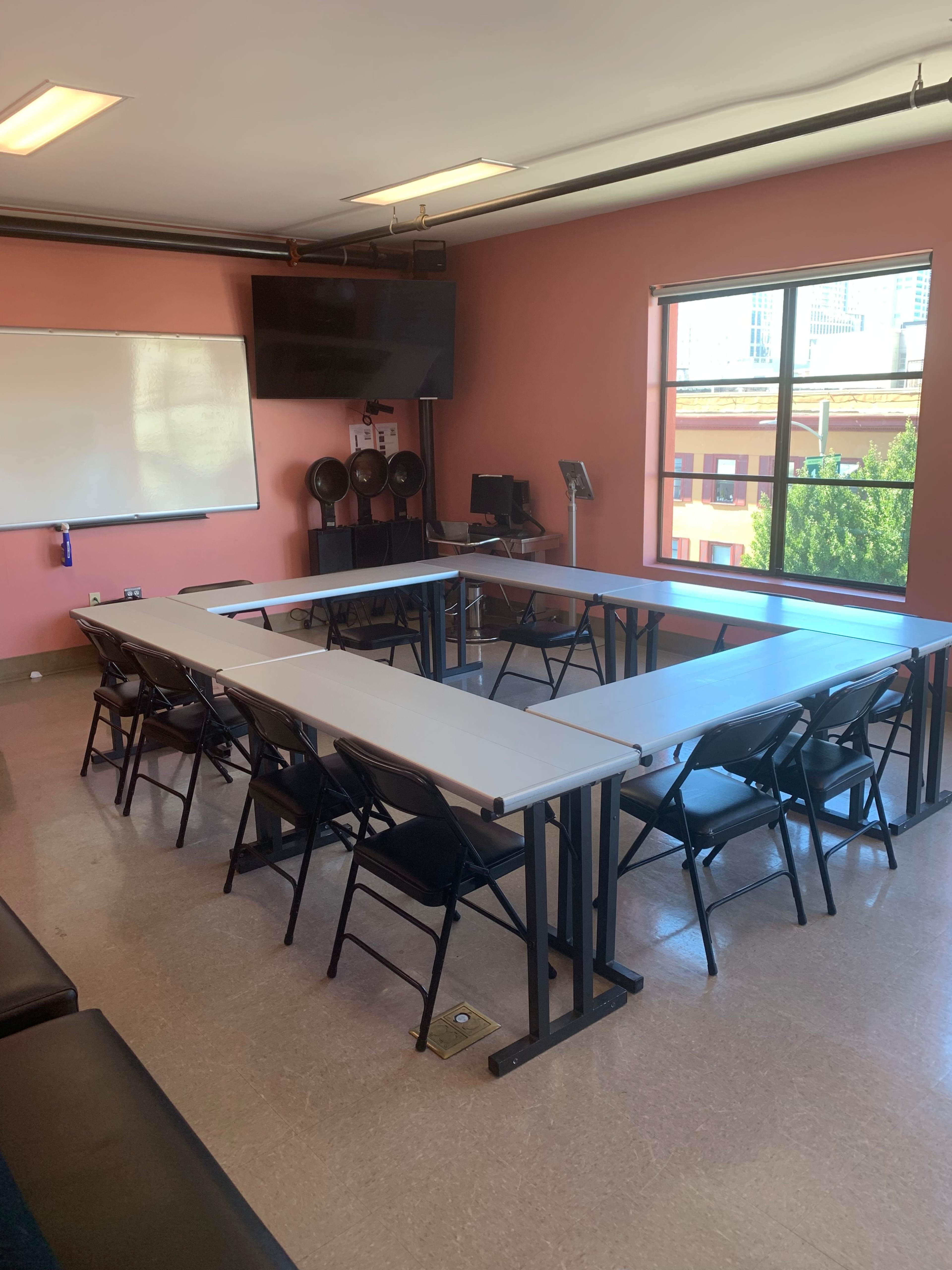A classroom features several tables arranged in a U-shape, with black chairs and a large window overlooking a cityscape.