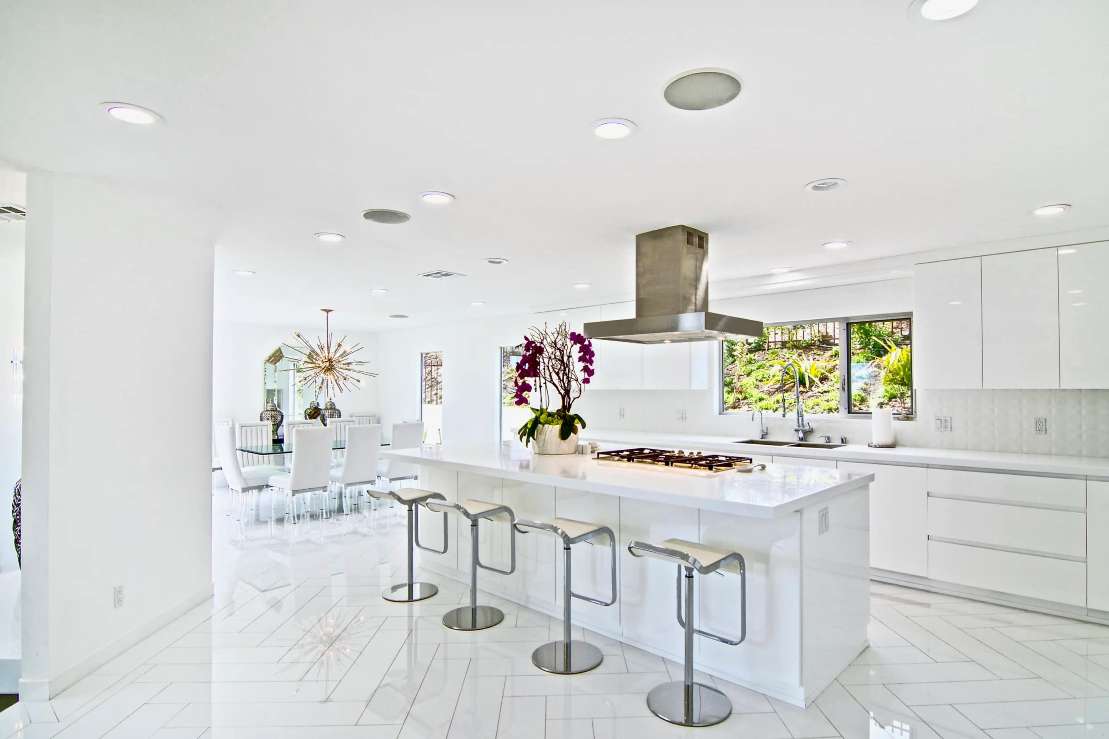 The image shows a modern kitchen with sleek white cabinetry, a large island with bar stools, and an adjoining dining area visible through large windows.