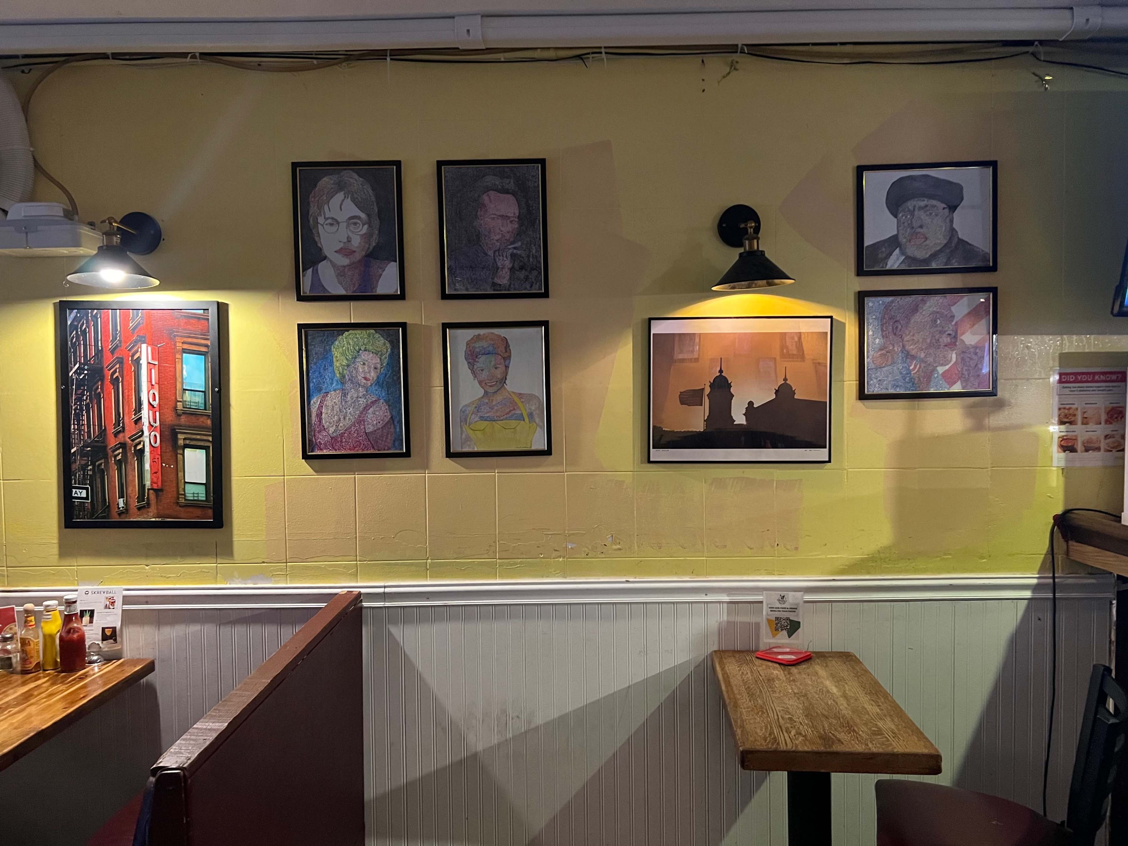 A wall in a restaurant adorned with various framed portraits and paintings, alongside a wooden table and seating area below.