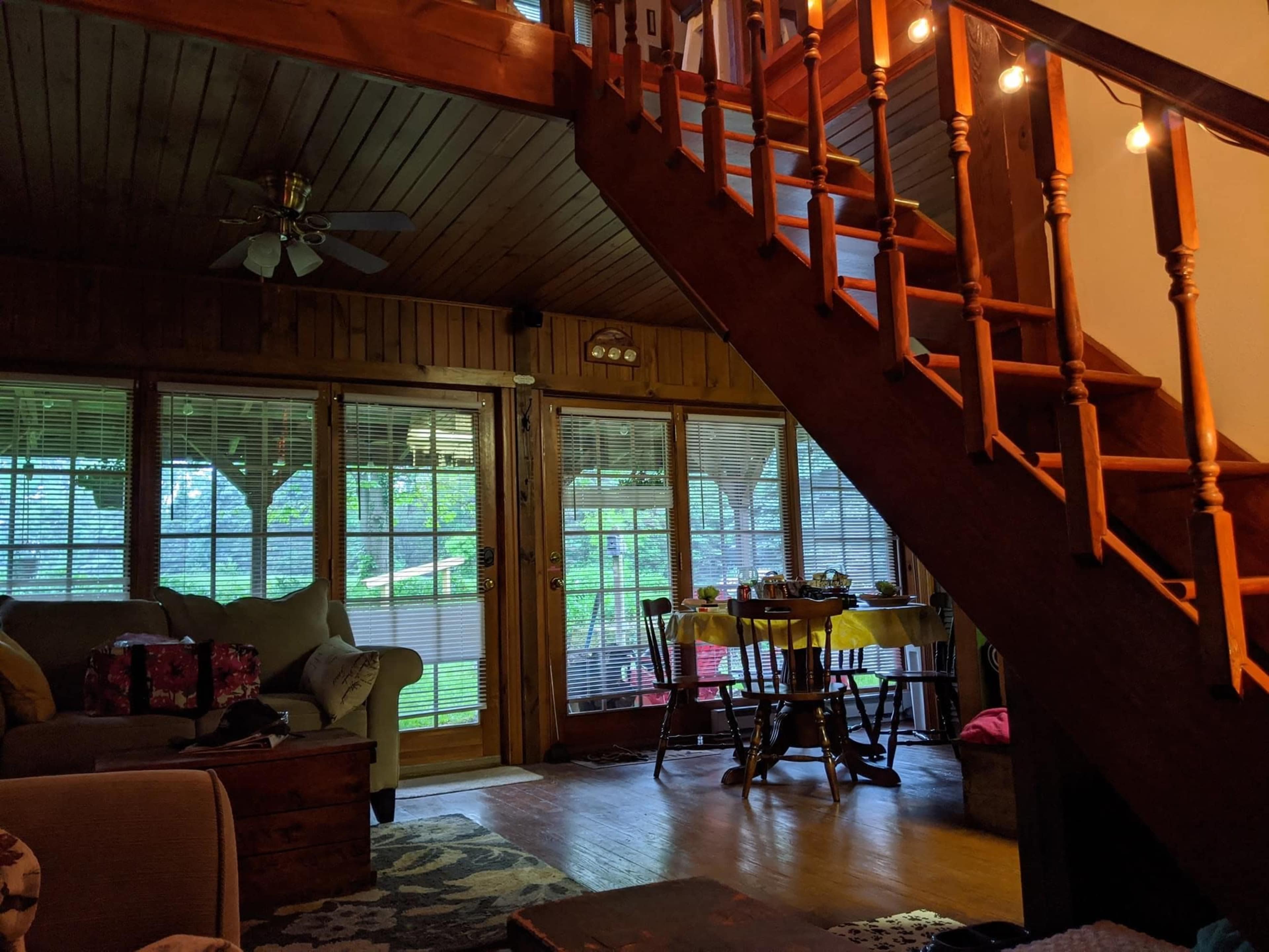 A cozy living area with a staircase leading to an upper level, large windows, a sofa, and a dining table set for a meal.