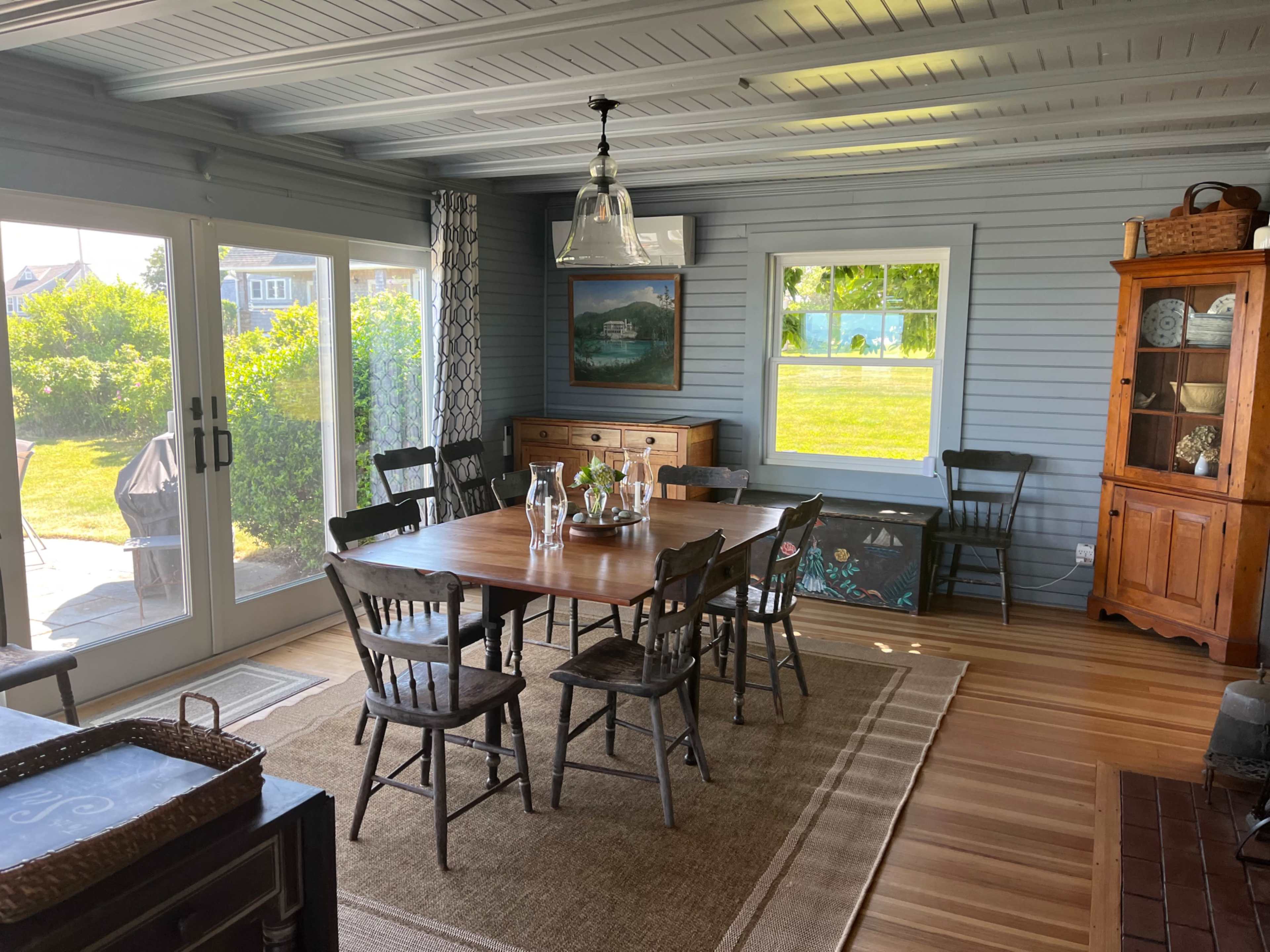 A sunlit dining area with a large wooden table surrounded by six black chairs, a sideboard, and a cupboard against the walls of a blue-paneled room.