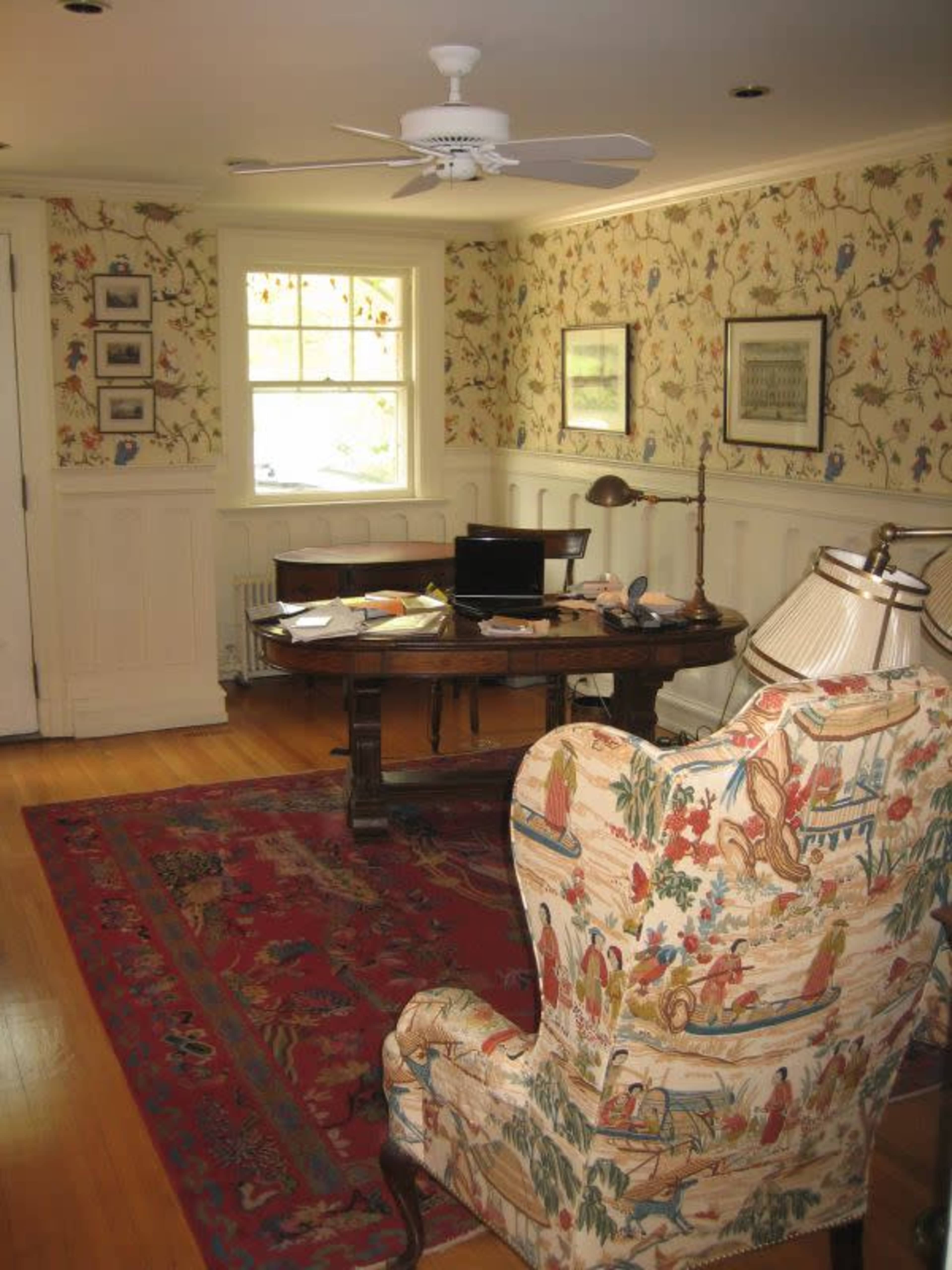 The image shows a well-lit home office with a wooden desk, a computer, and a large upholstered chair beside a patterned area rug.