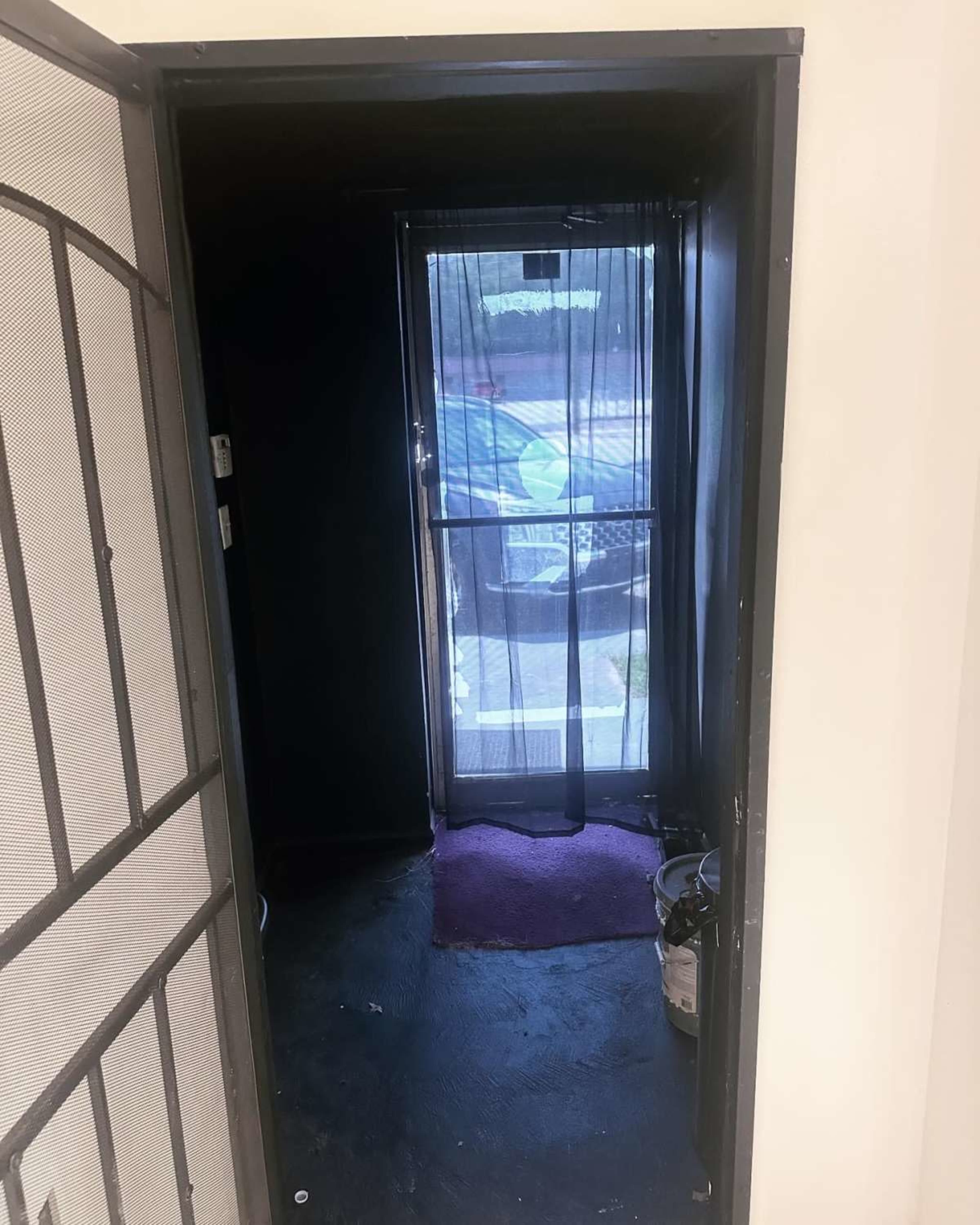The image shows a doorway with a sheer curtain leading to an outdoor area, featuring a dark interior and a small purple mat on the floor.