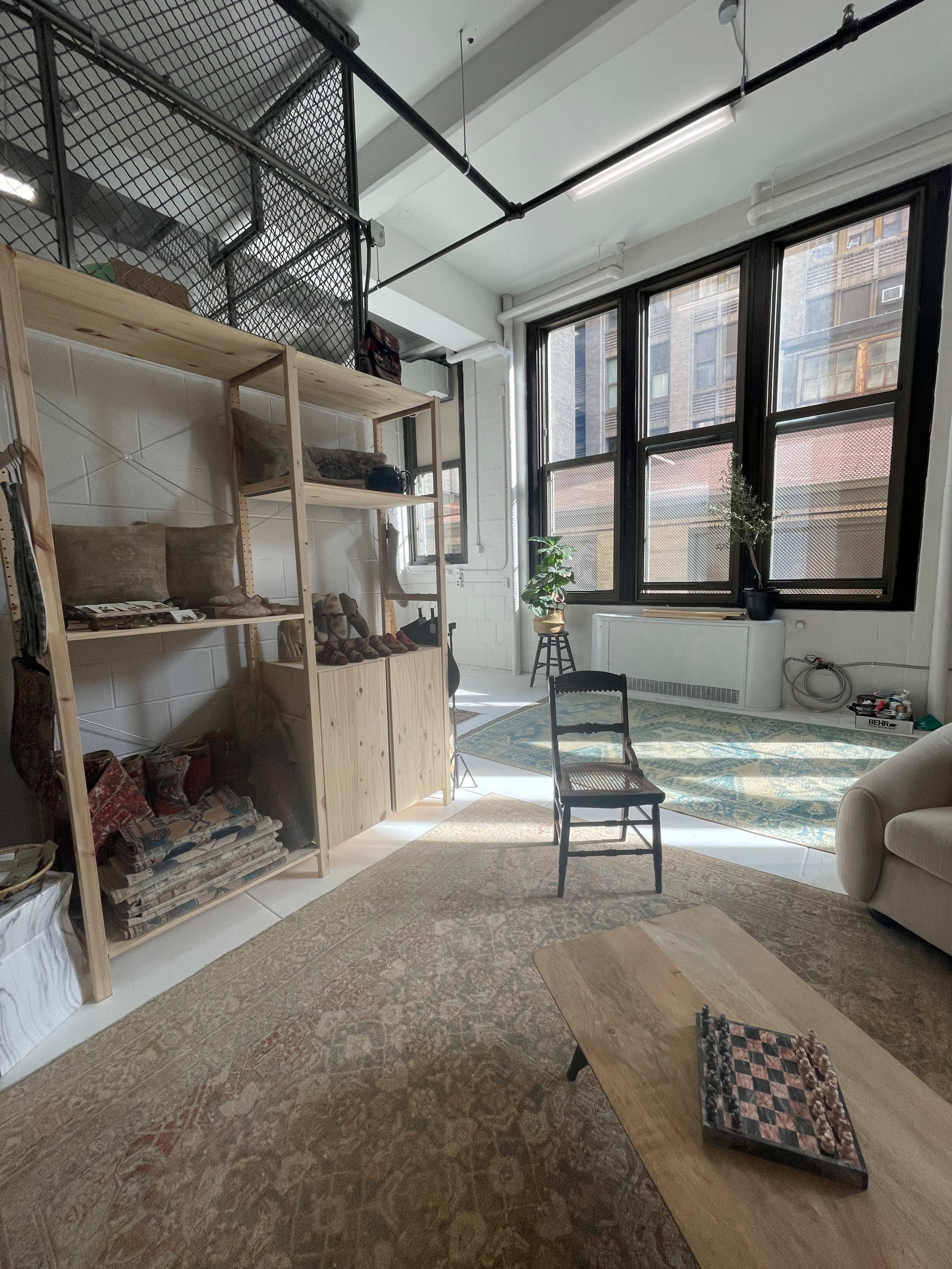 A bright, minimalist room with large windows, wooden shelving filled with items, a chair, and a comfortable sofa arranged on a patterned rug.
