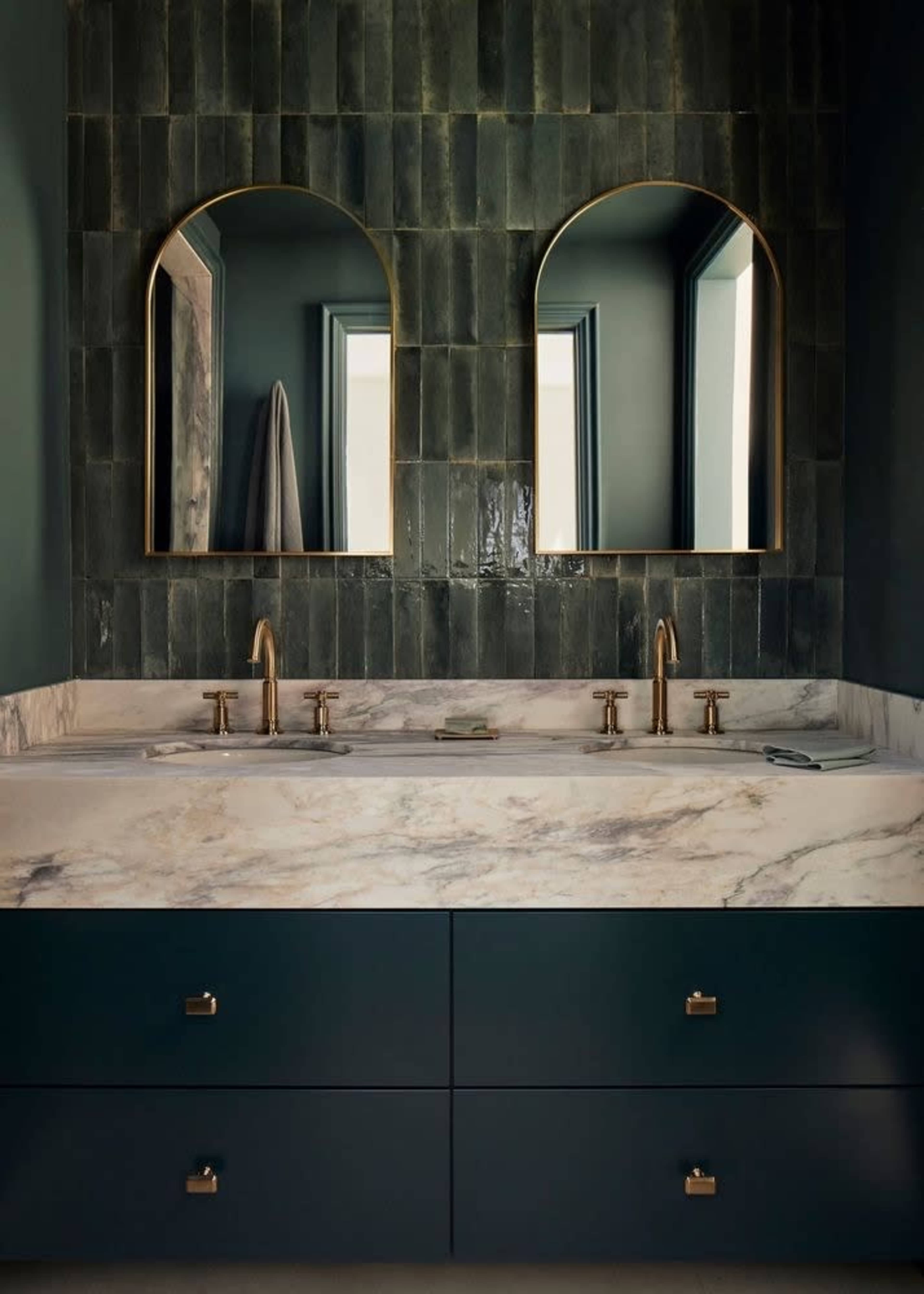 The image shows a modern bathroom with a double sink vanity featuring a marble countertop, dark green tile walls, and two arched mirrors framed in gold.