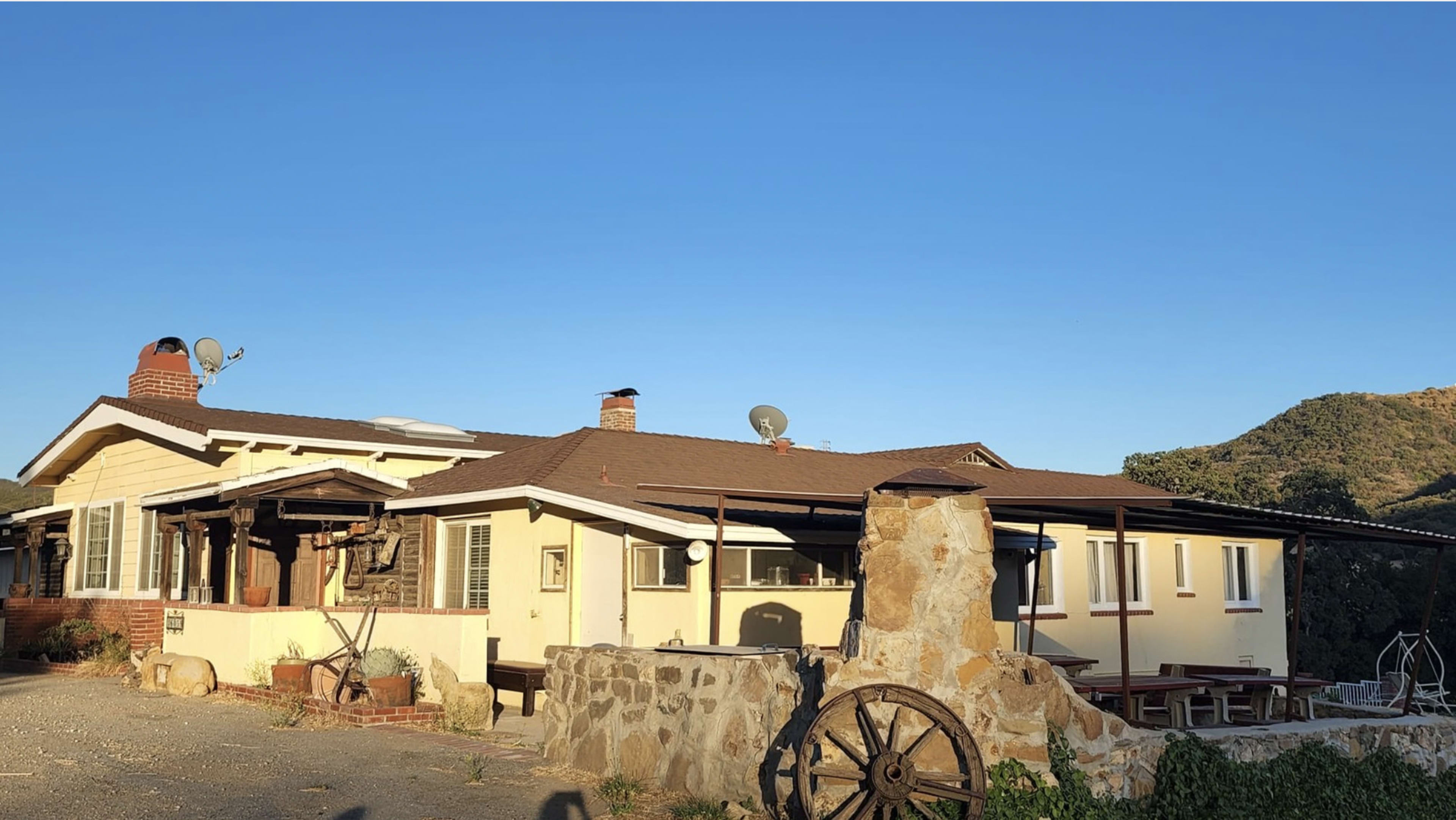A yellow rural house with a stone wall and an old wooden cart wheel is set against a clear blue sky and hills in the background.
