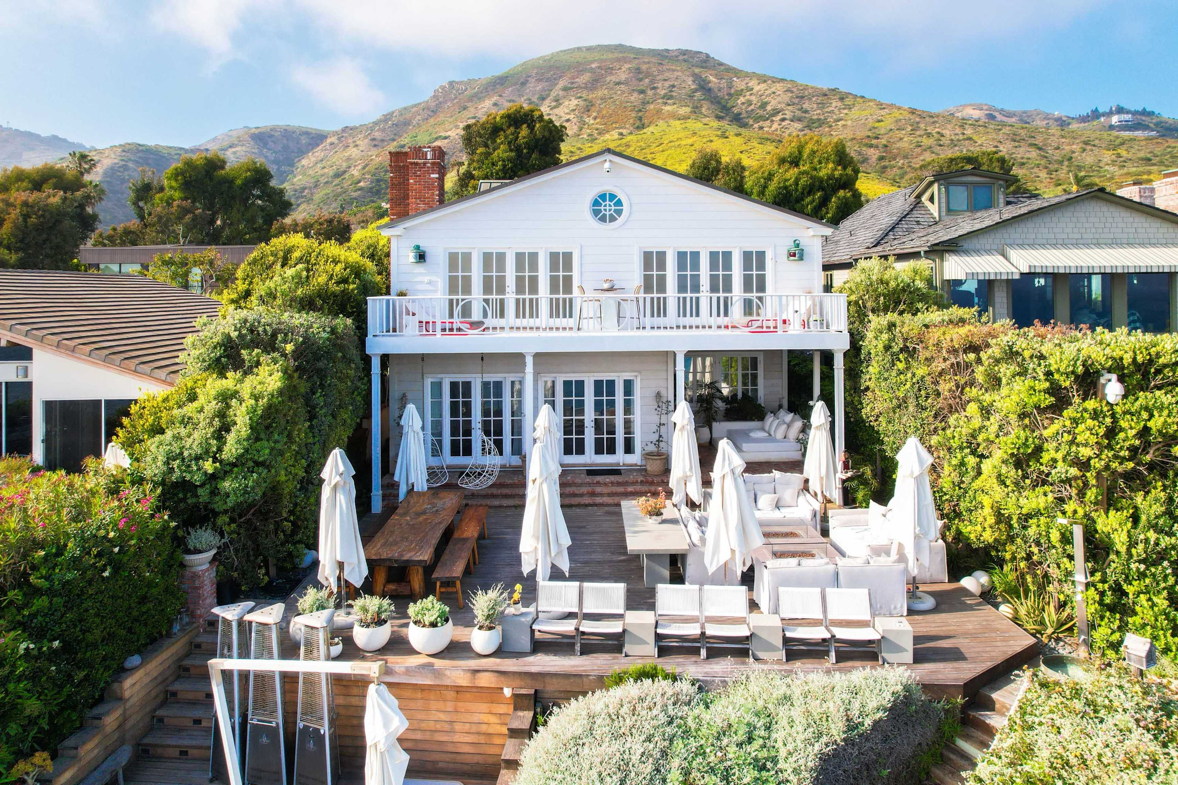 The image shows a two-story white house surrounded by greenery, with a spacious outdoor patio featuring lounge chairs and dining areas, set against a backdrop of a hillside.