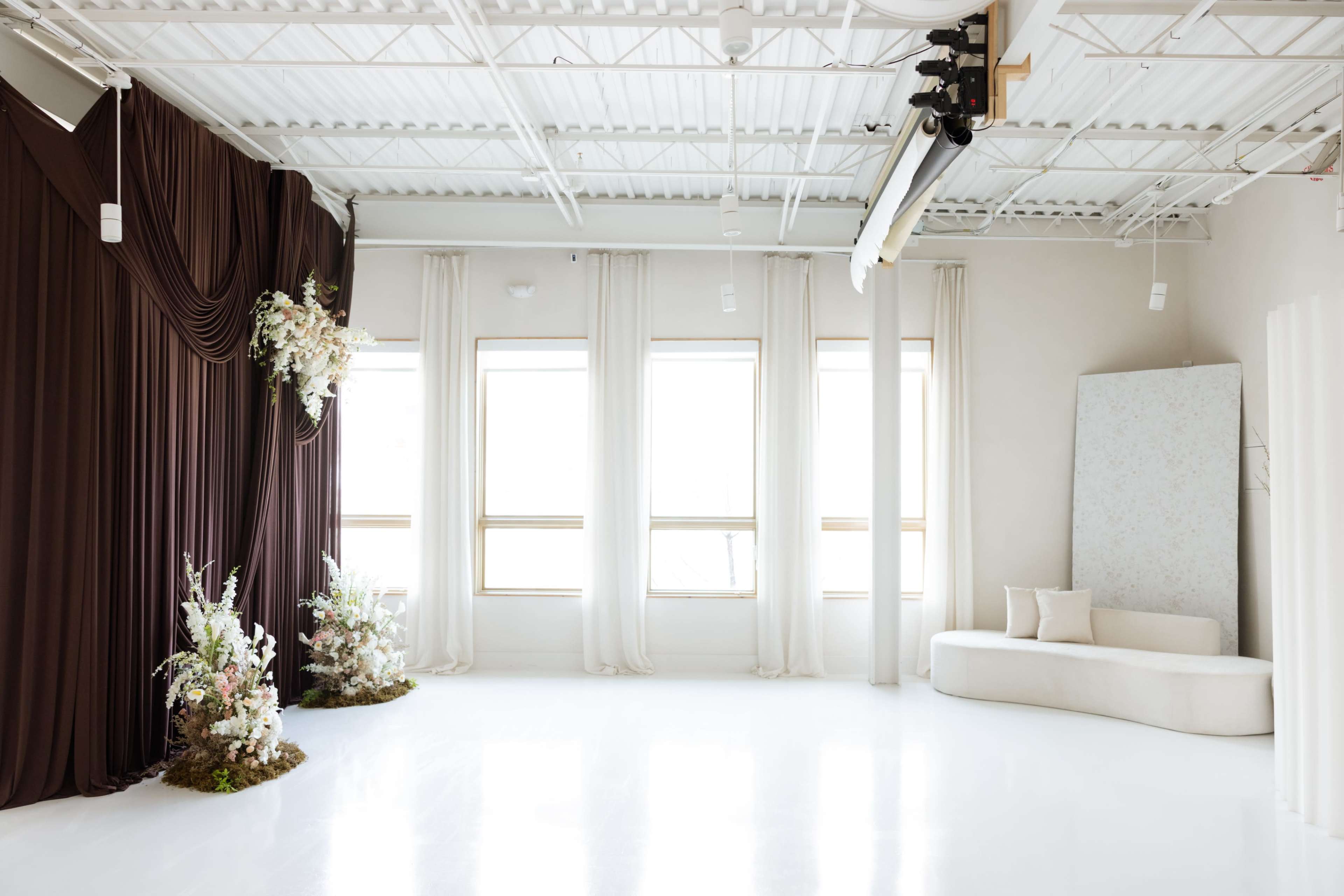 The image features a spacious studio interior with large windows, white flooring, a curved sofa, and decorative fabric drapes on one side.