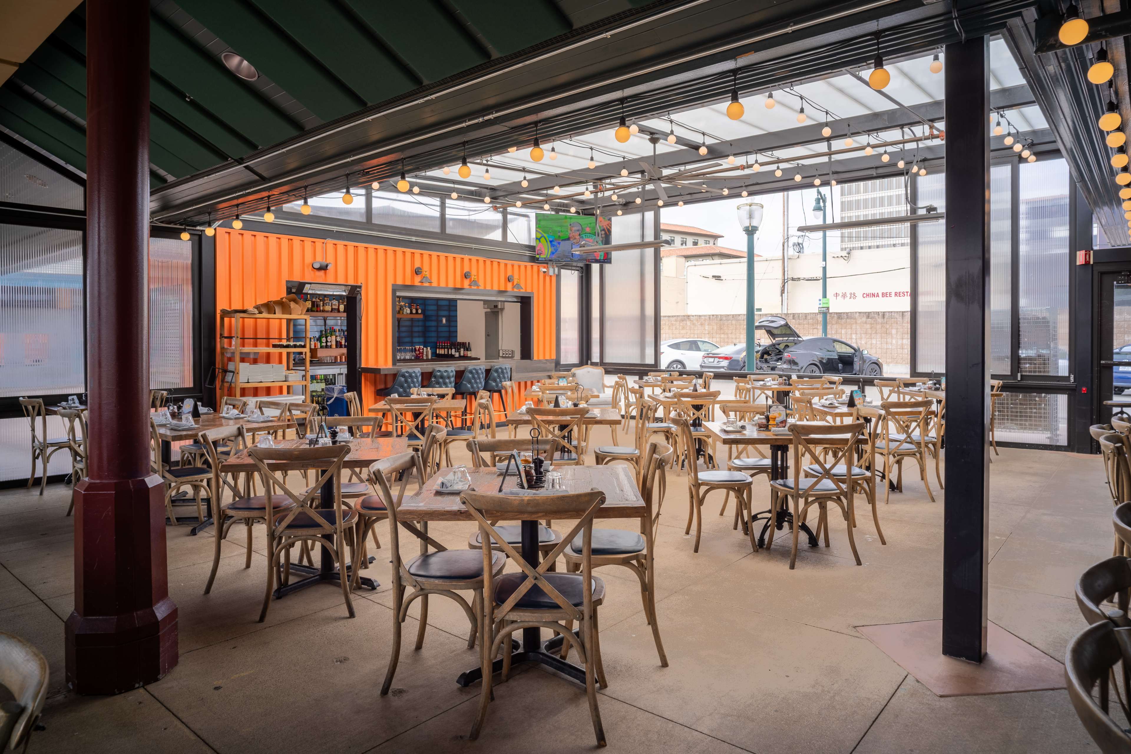The image shows an empty restaurant patio with rows of wooden tables and chairs, illuminated by string lights overhead.