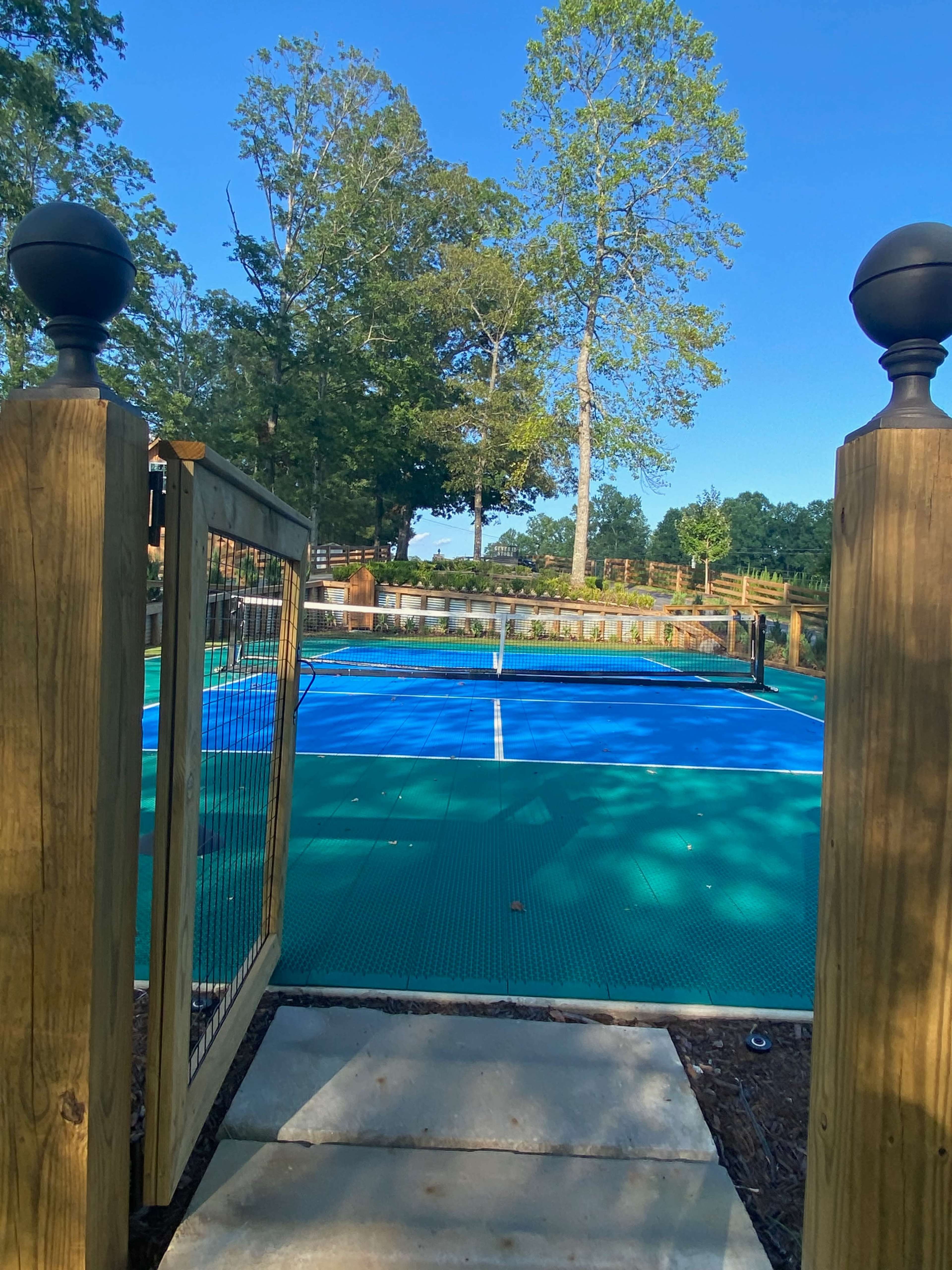 A gated entrance leading to a blue tennis court surrounded by trees.