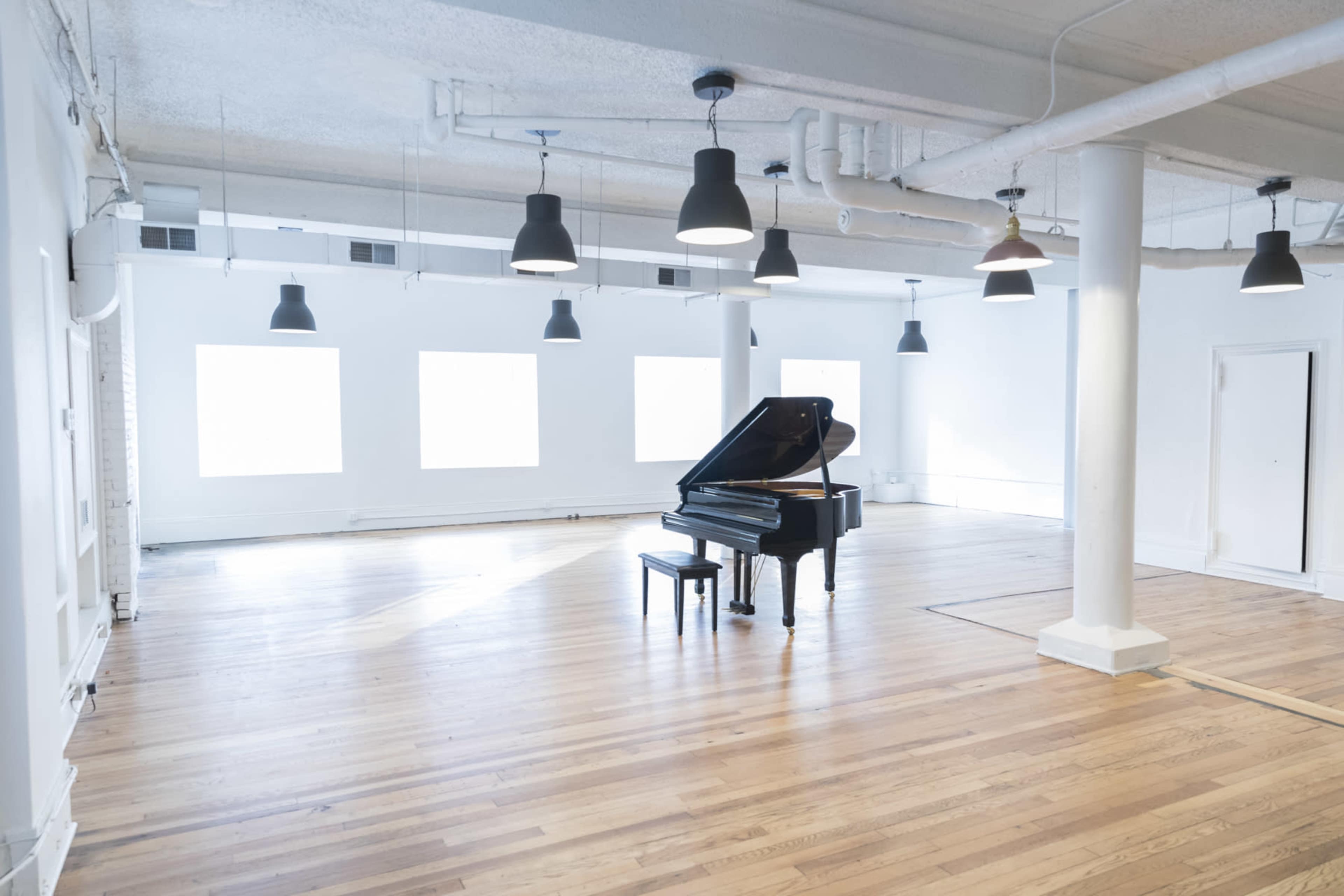 A grand piano sits alone in a spacious, brightly lit room with wooden floors and large windows.