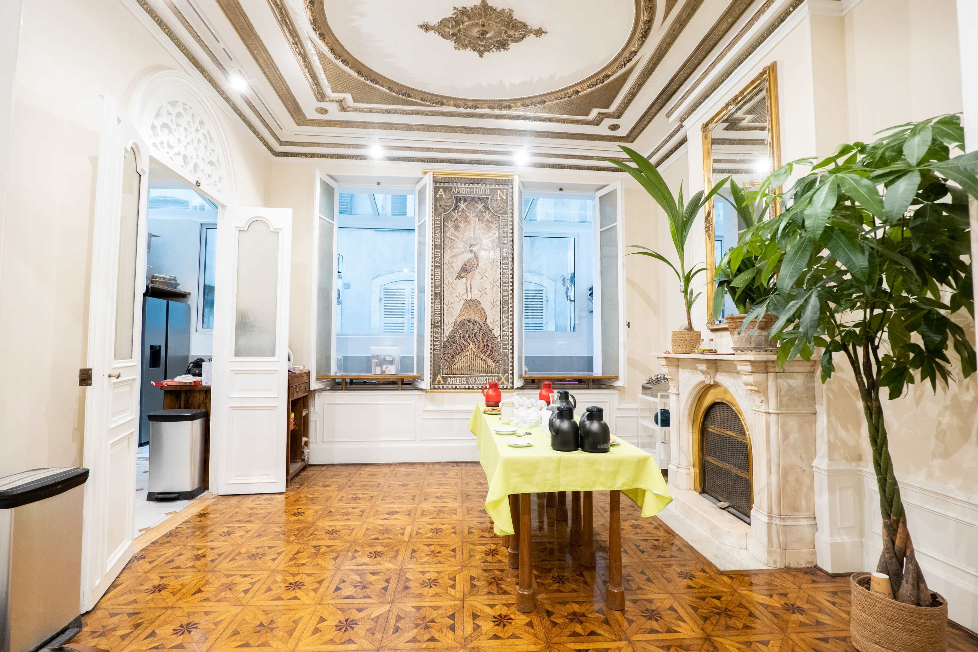 The image shows a spacious room with a decorative ceiling, two large windows, and a table set with dining items in the center.