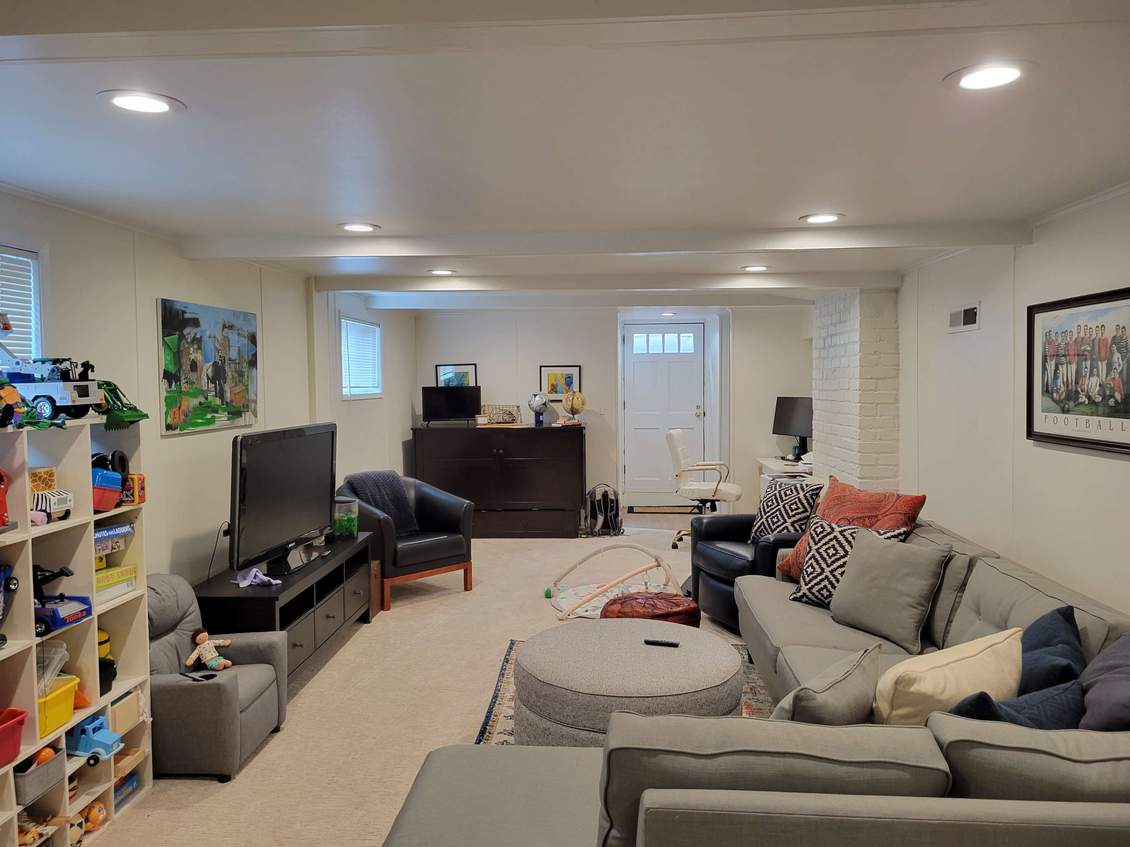 The image shows a cozy basement living area with a sectional sofa, a coffee table, a TV stand, and various toys organized on shelves.