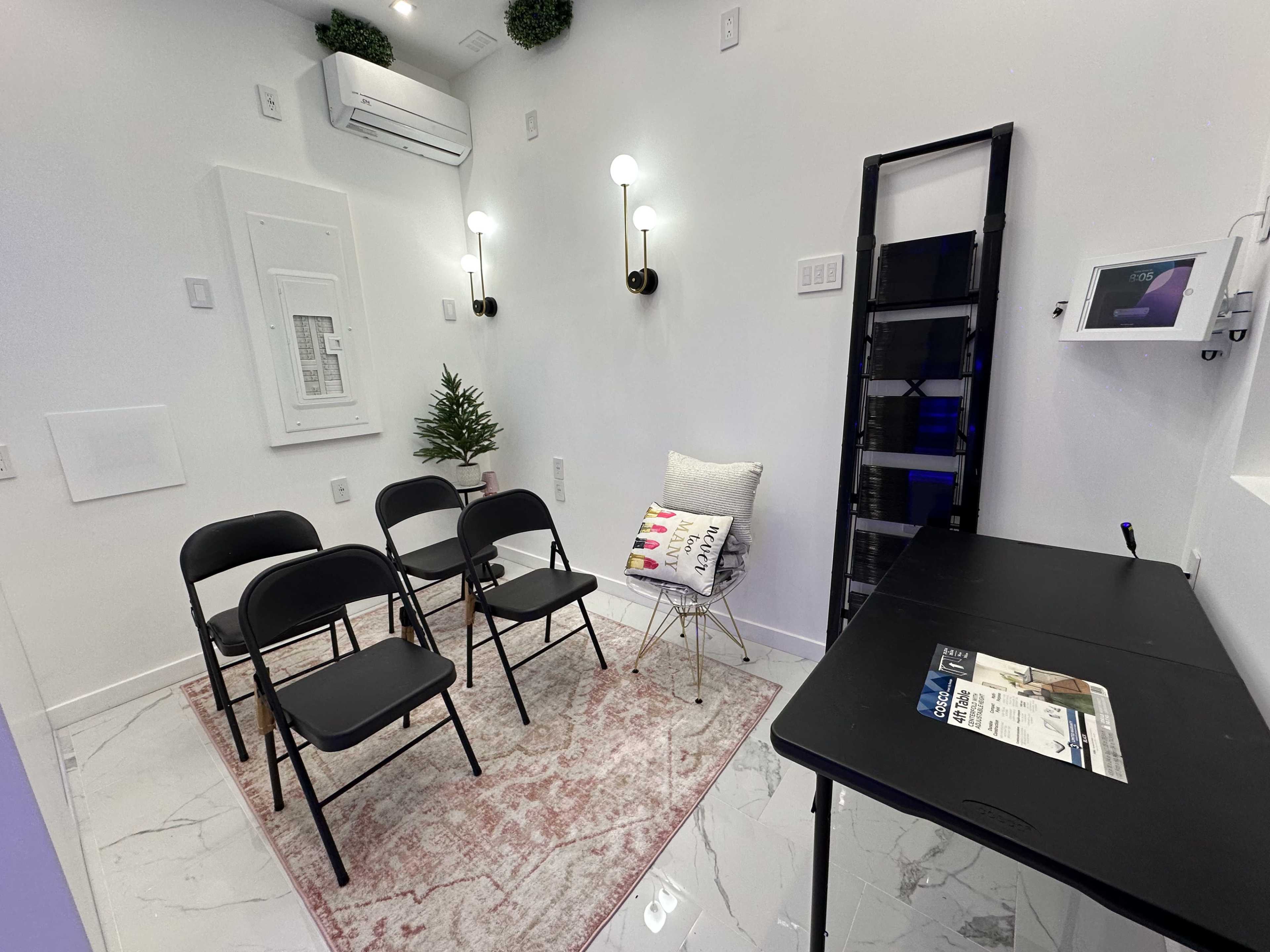 The image shows a small, minimalist room with four black chairs arranged in front of a white wall, a small plant, and a side table with a magazine.