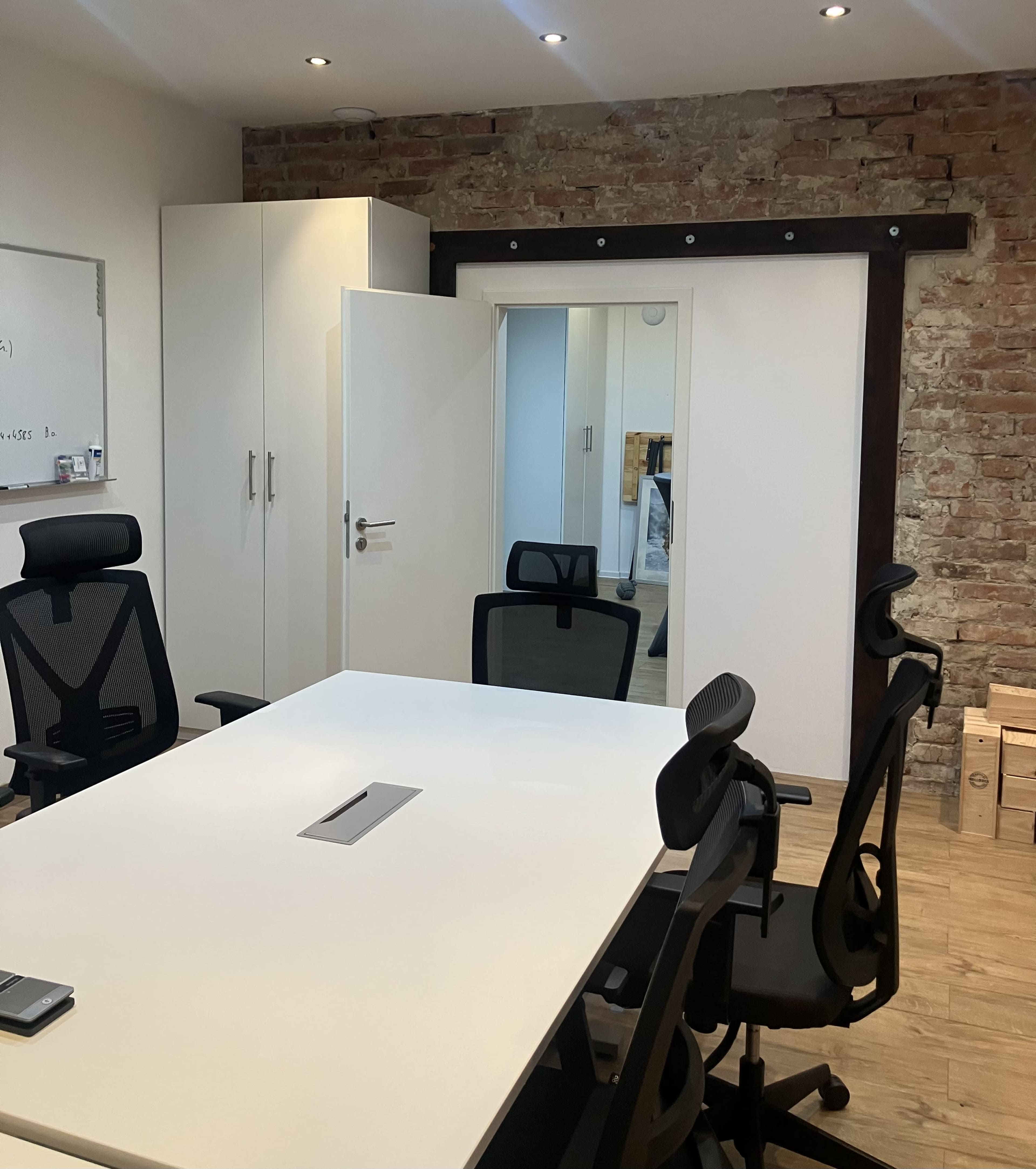 The image shows a modern meeting room with a white table surrounded by black office chairs, a sliding door, and a brick wall in the background.