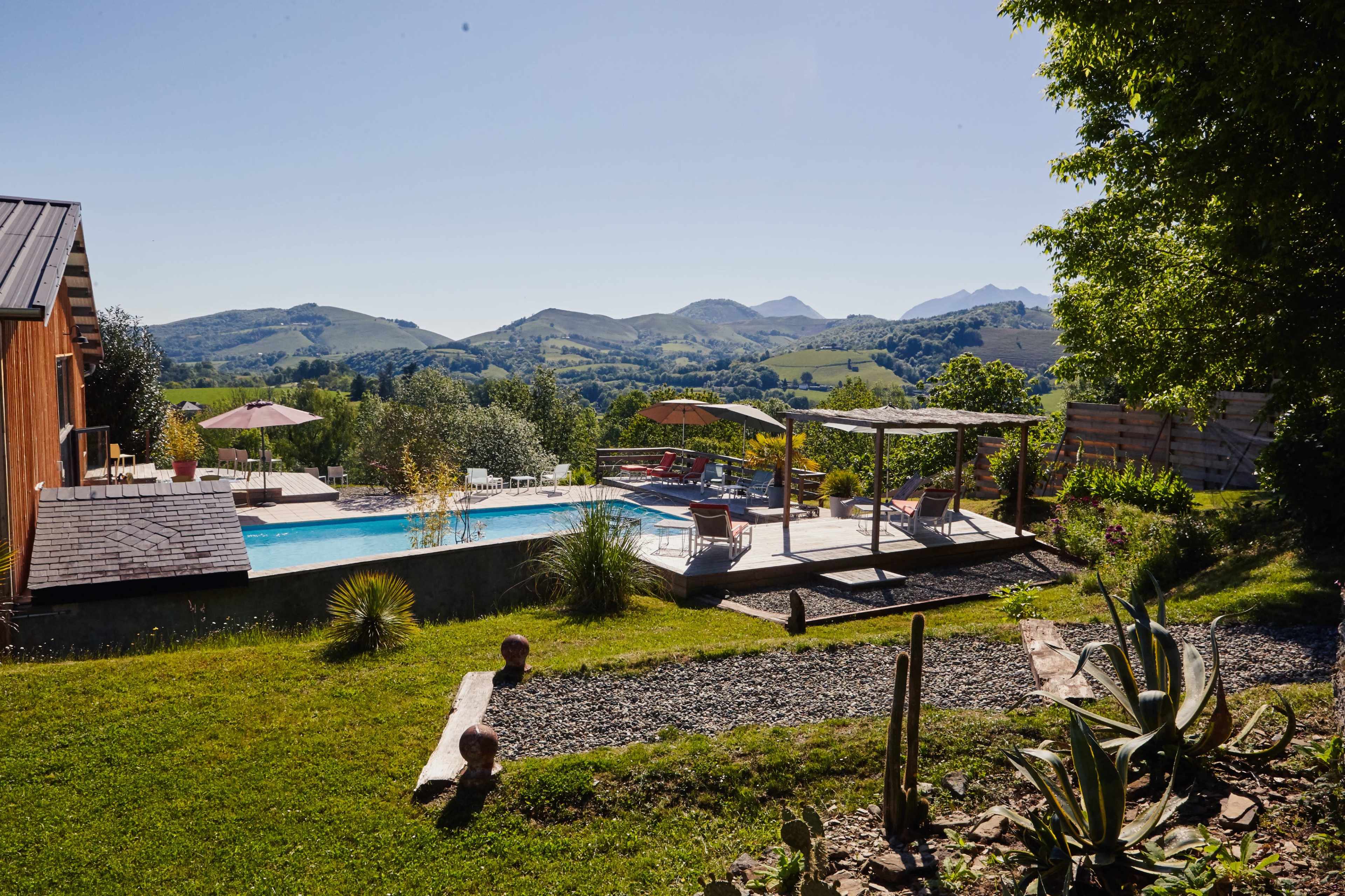 A landscaped backyard features a swimming pool, lounge area, and seating under a pergola with mountains in the background.