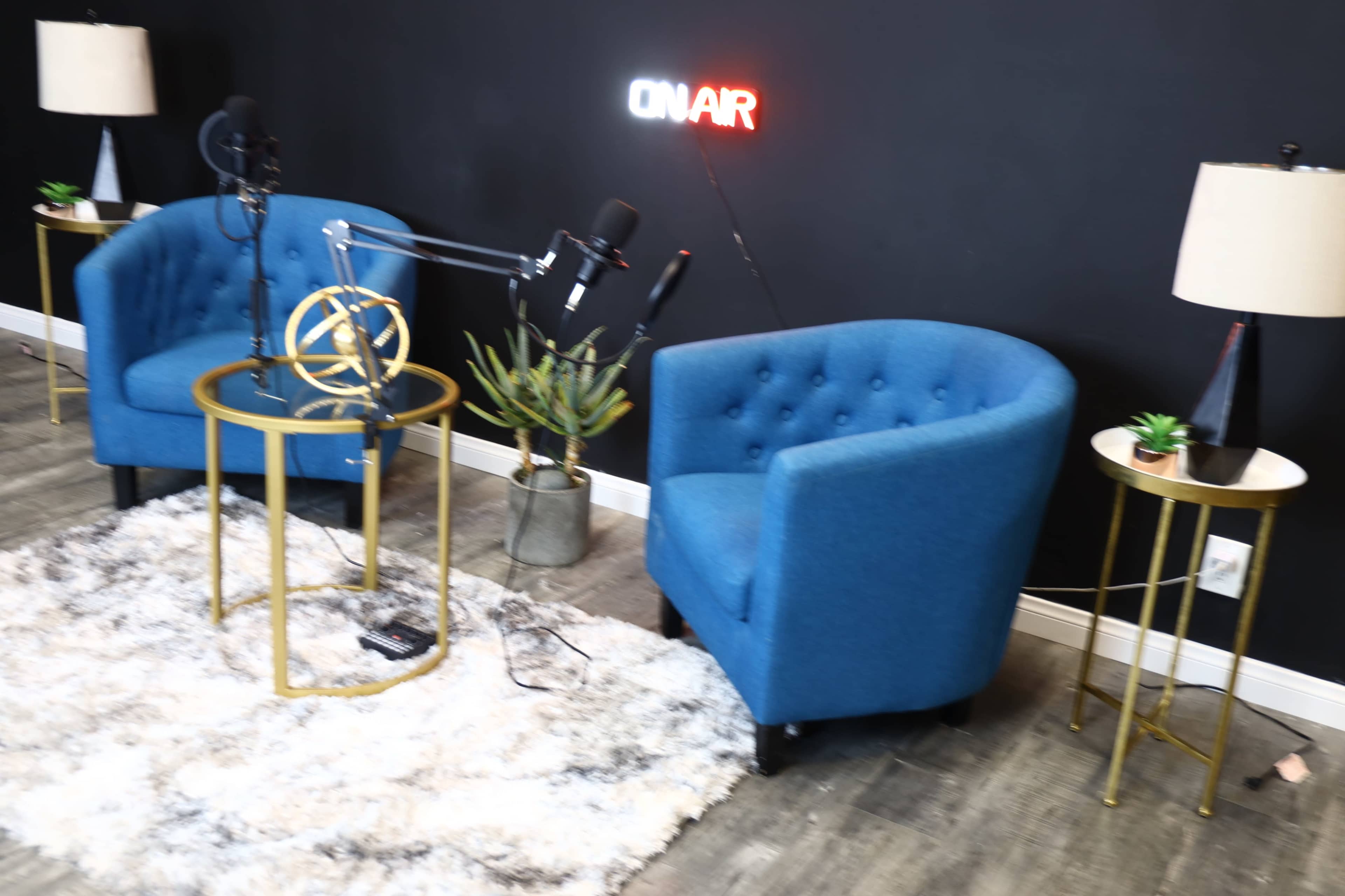 The image shows a podcast studio setup featuring two blue chairs, a round glass table with a decorative globe, and a wall-mounted "ON AIR" sign.