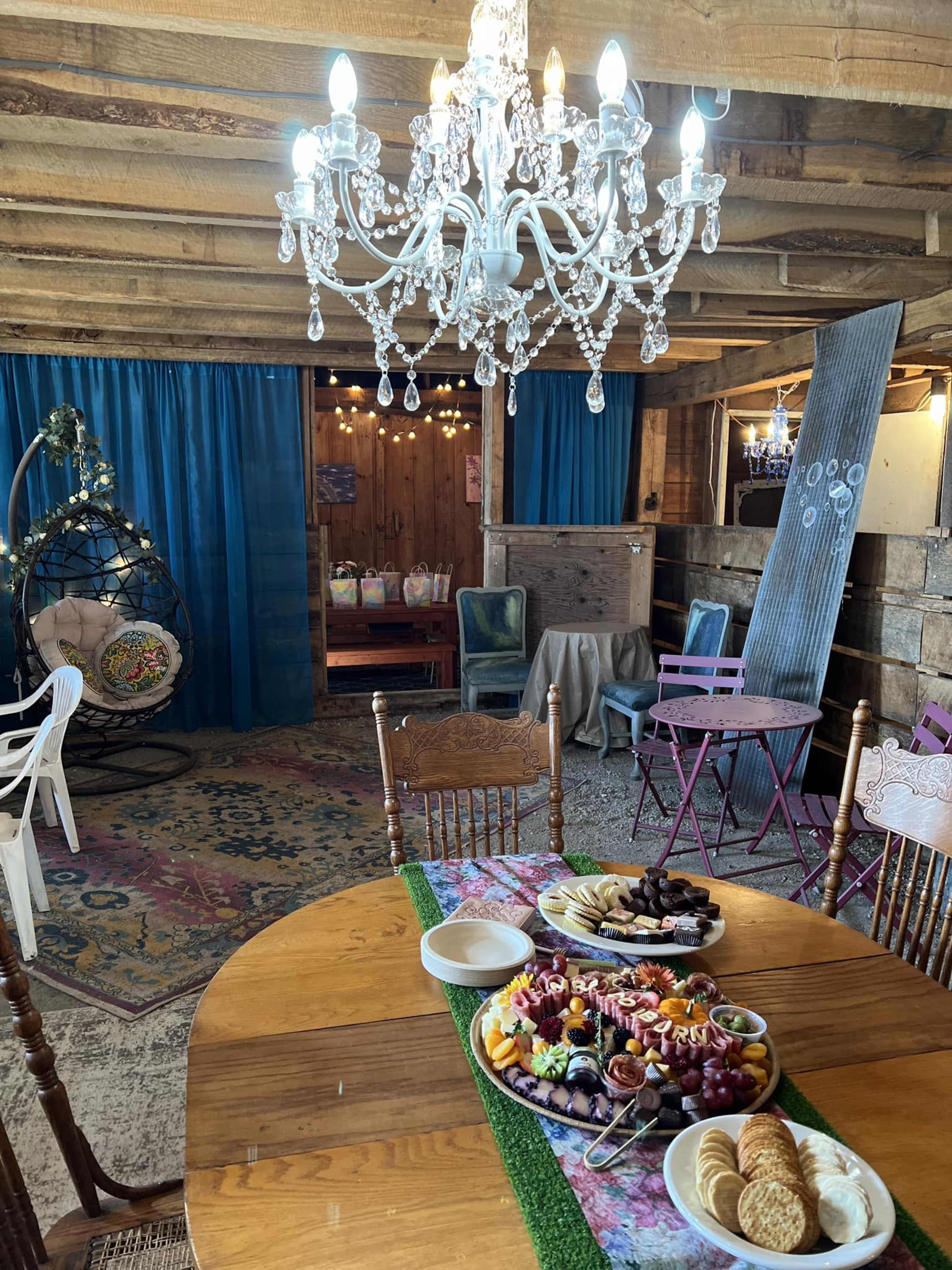 A dining table is set with a variety of food, including a fruit platter, in a rustic room featuring a chandelier and blue curtains.