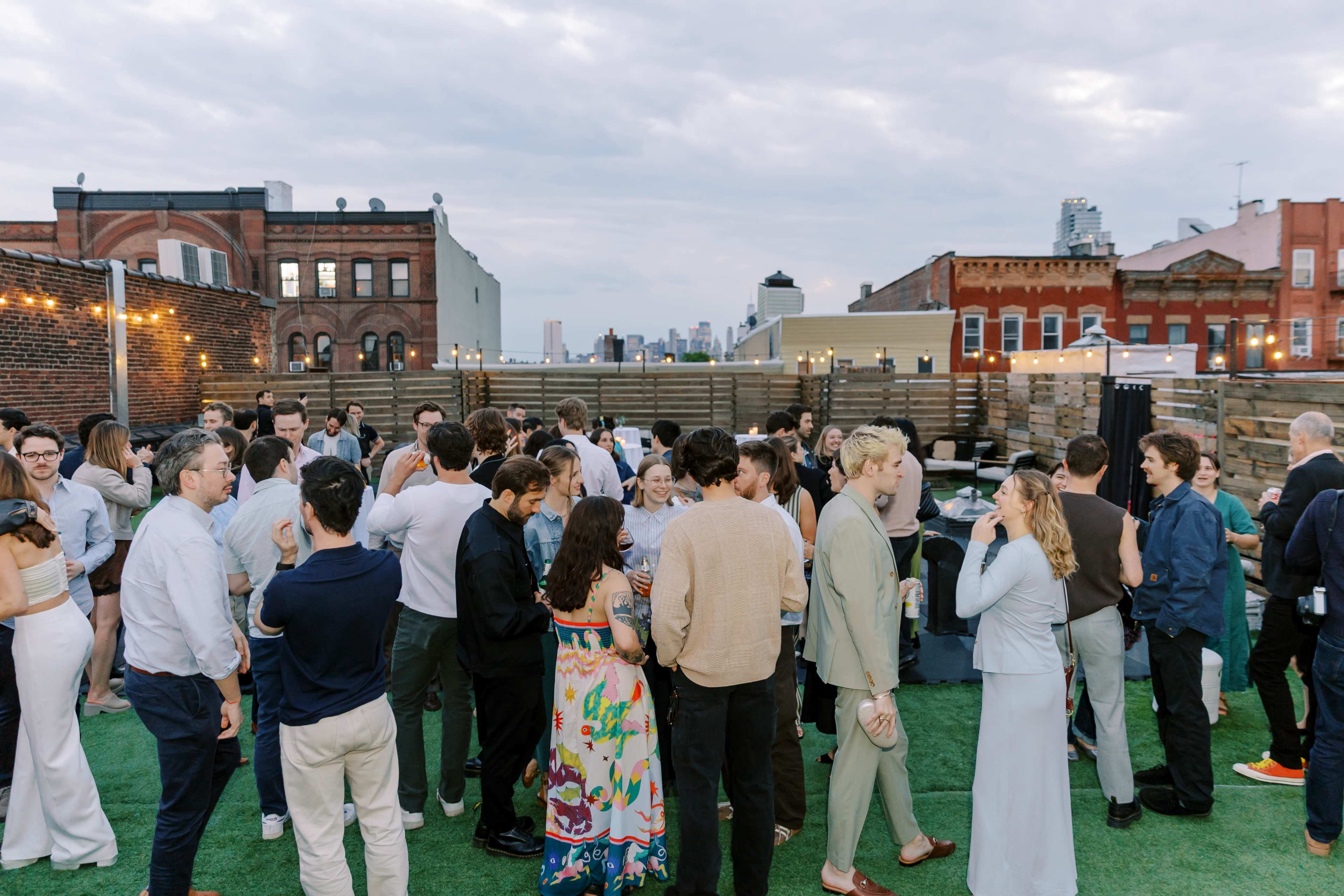 A large group of people mingles on a rooftop terrace, surrounded by city buildings under a cloudy sky.