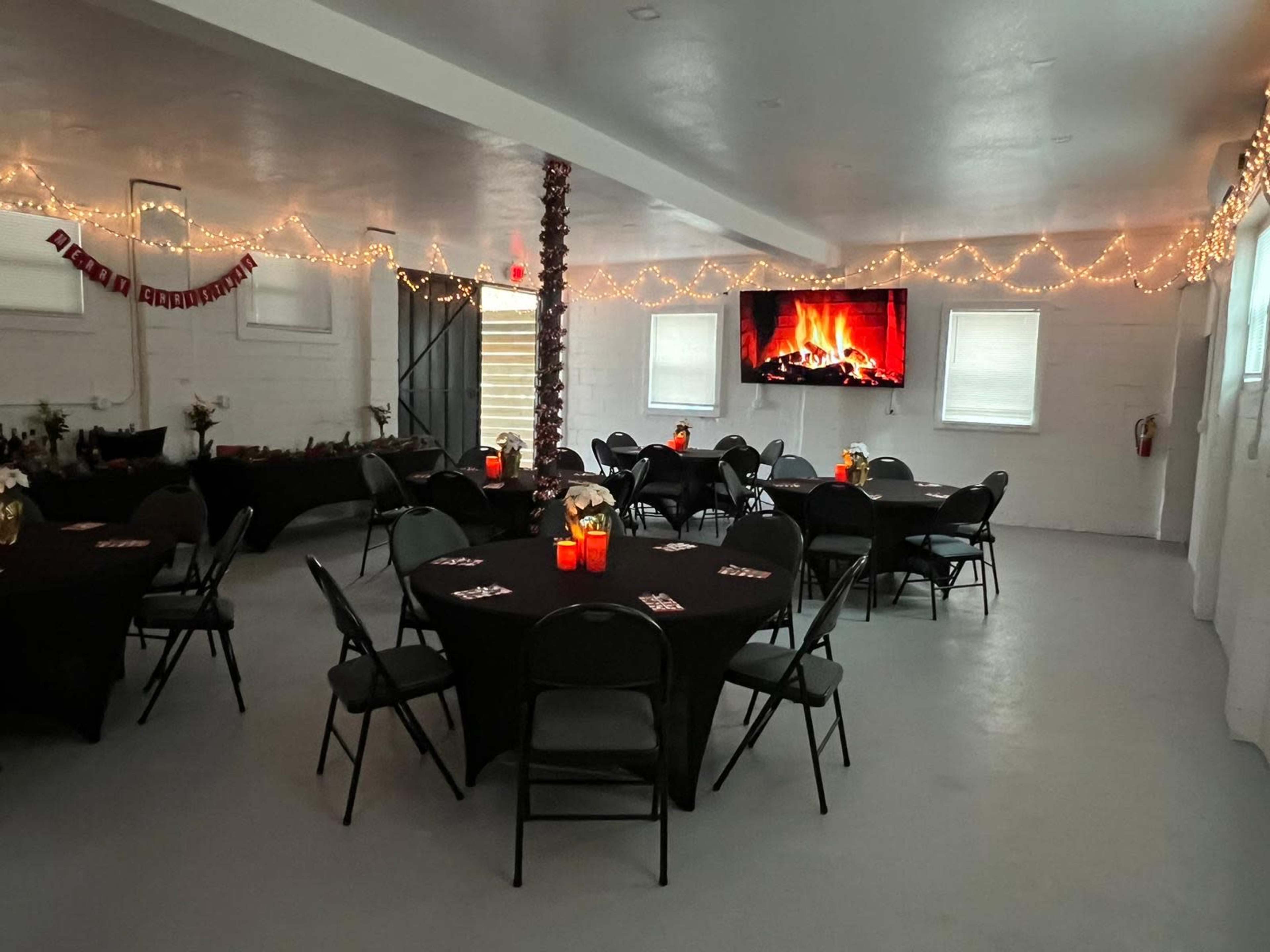 A decorated indoor event space features black tables with white tablecloths, chairs arranged around them, and a large screen displaying a fireplace, accompanied by string lights and festive decor.