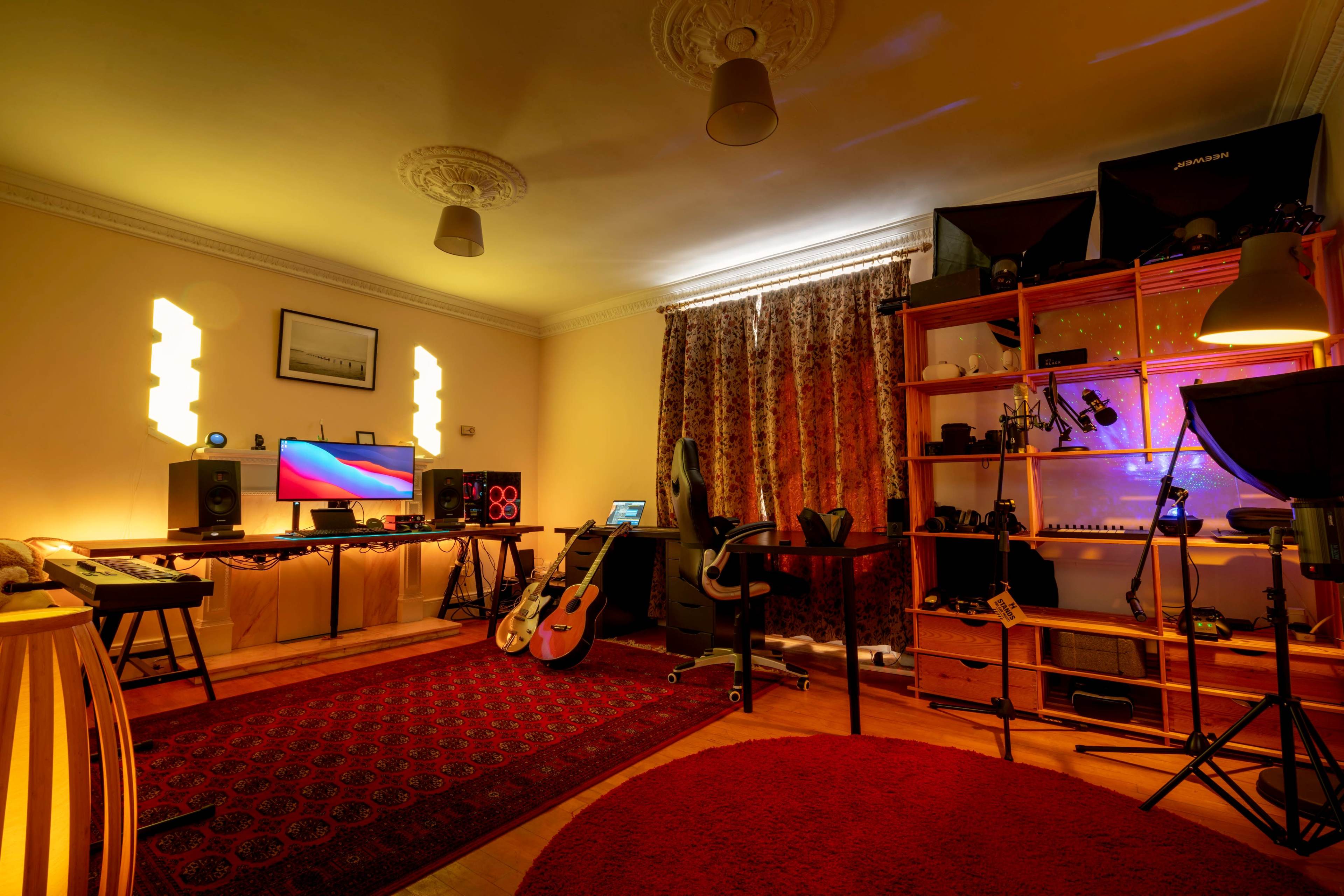 A music production studio featuring two computer monitors, musical instruments, and various equipment arranged around a cozy room with patterned curtains and a circular rug.
