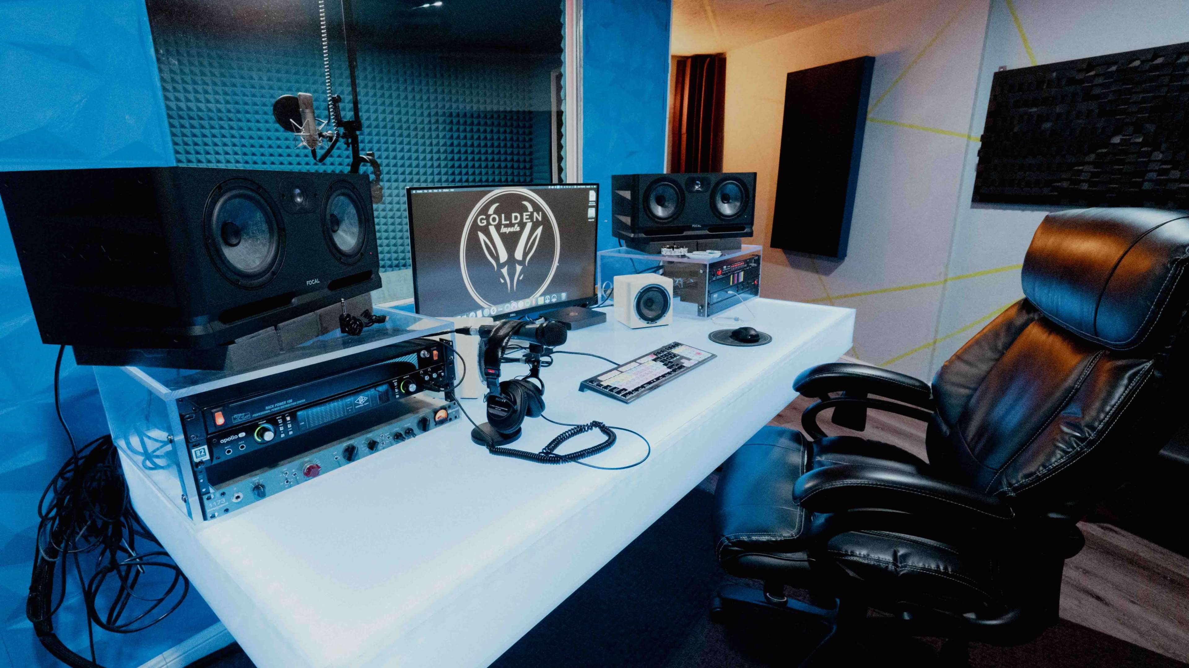 The image shows a recording studio setup featuring a desk with speakers, a computer monitor displaying a logo, and audio equipment.