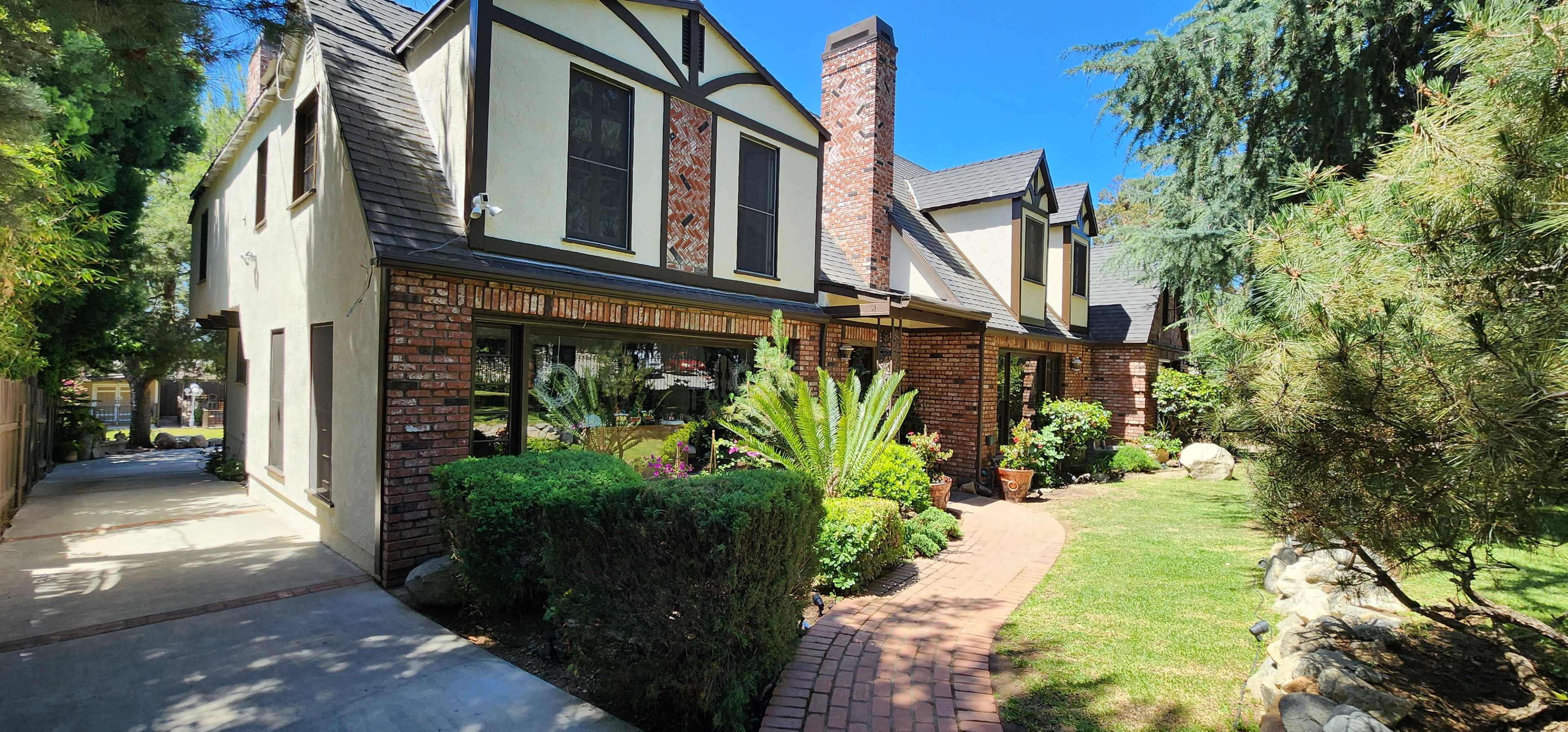 The image shows a brick house with a sloped roof, flanked by greenery and a walkway made of bricks, under a clear blue sky.