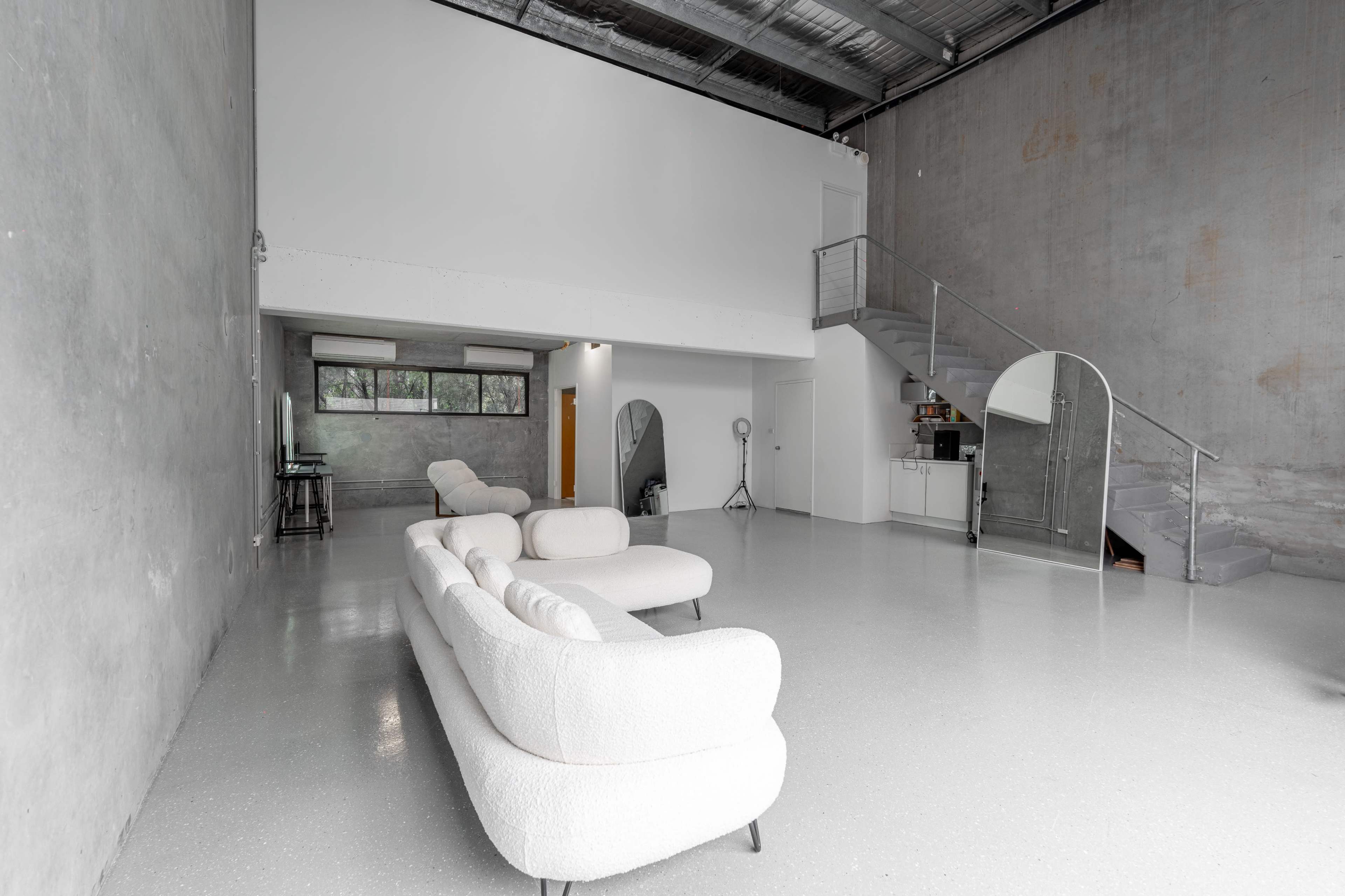 A spacious, minimalist interior featuring a white, curved sofa, large windows, and a modern staircase against a concrete wall.