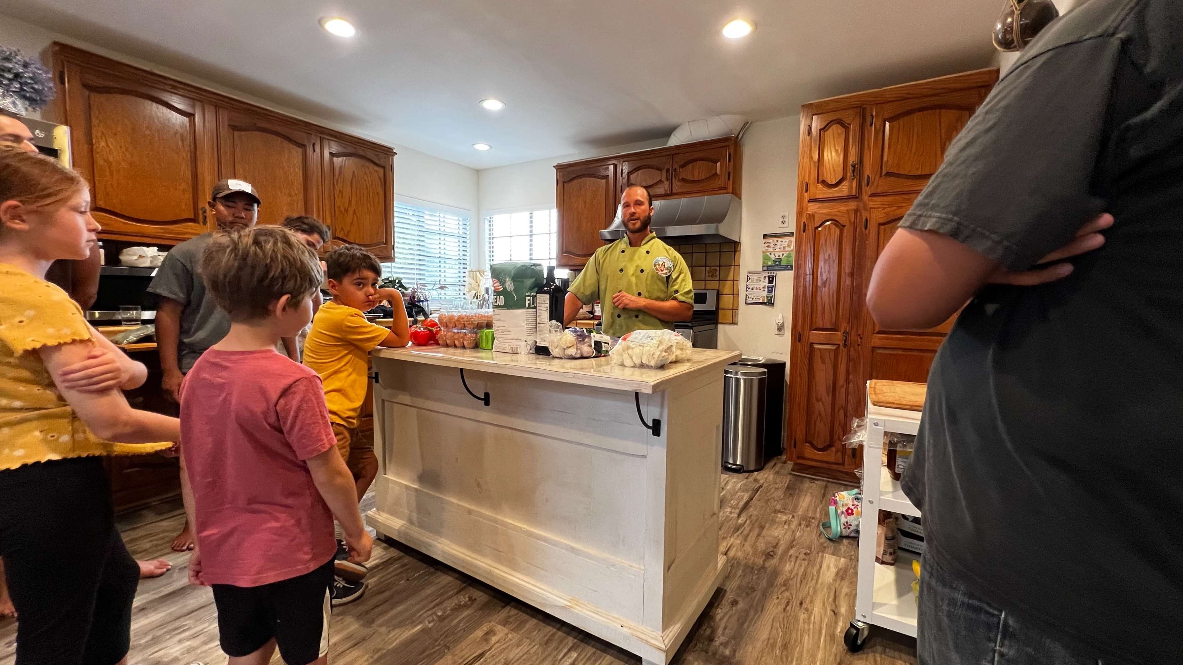 A group of people gathers around a kitchen island while a man explains something during a cooking demonstration.