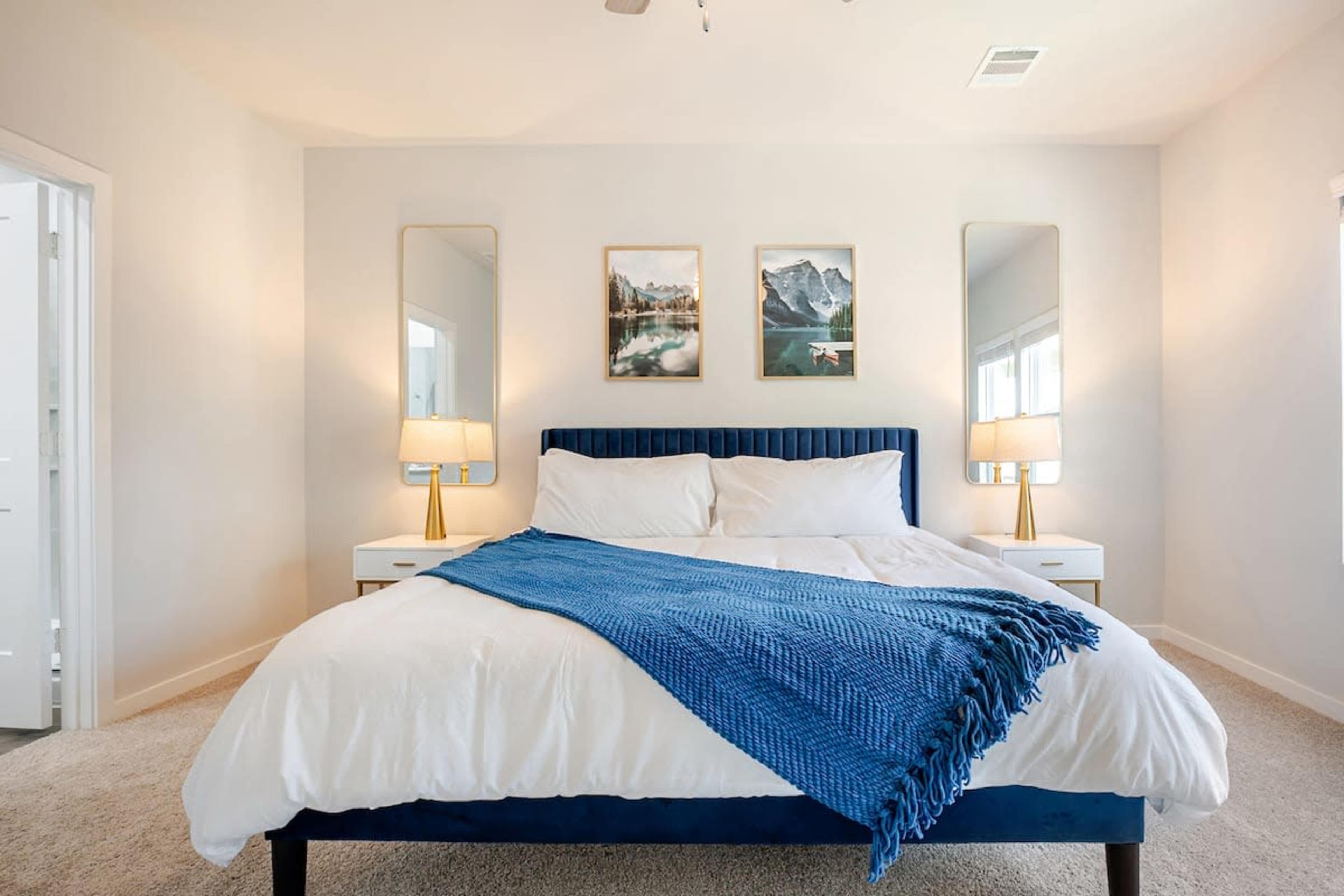 A large bed with a blue throw blanket is centered in a bright, minimalist bedroom featuring two mirrors and framed landscape art on the walls.