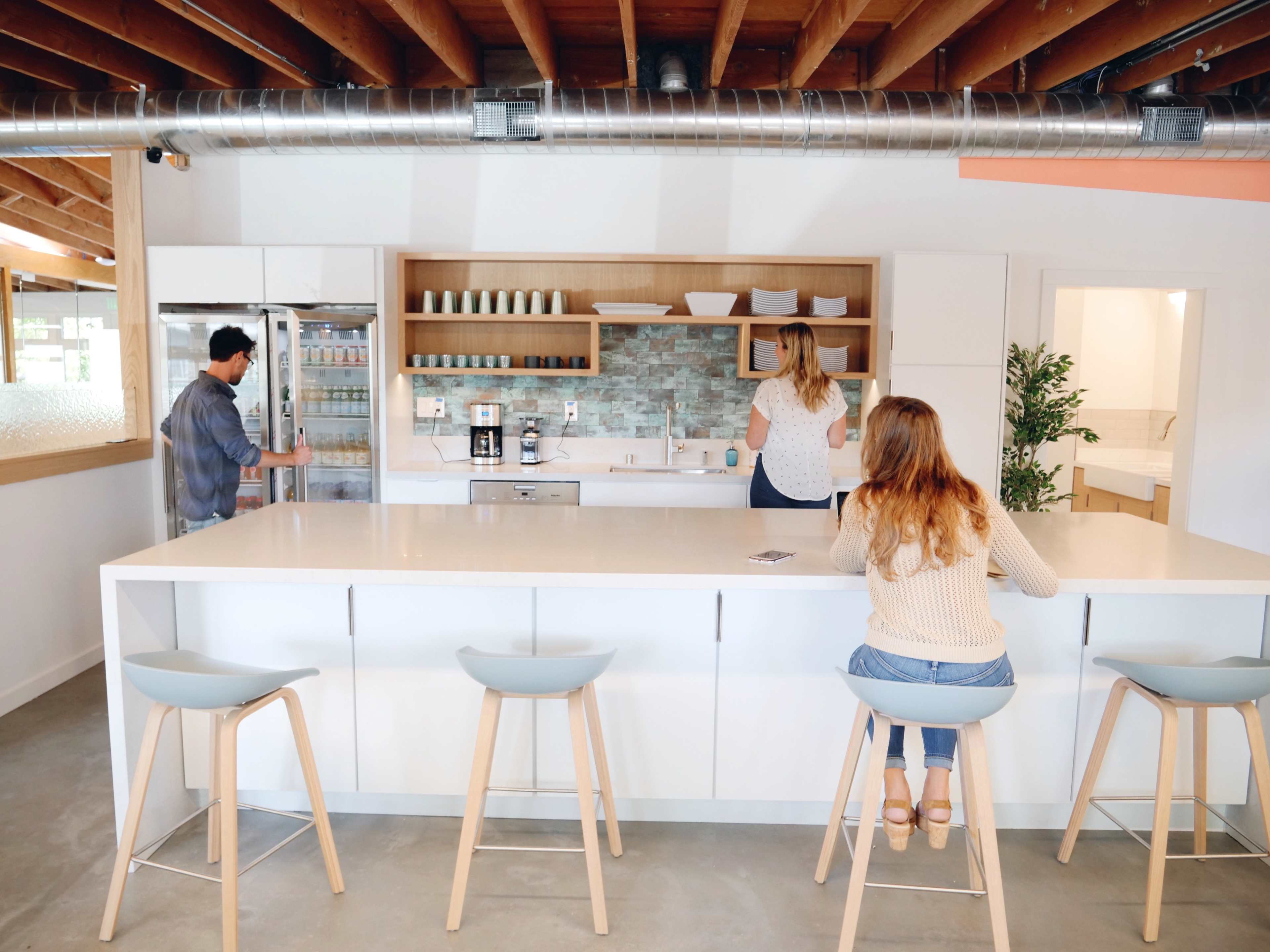 A modern kitchen features a large white island with stools, where one person sits while two others interact near a refrigerator and sink.