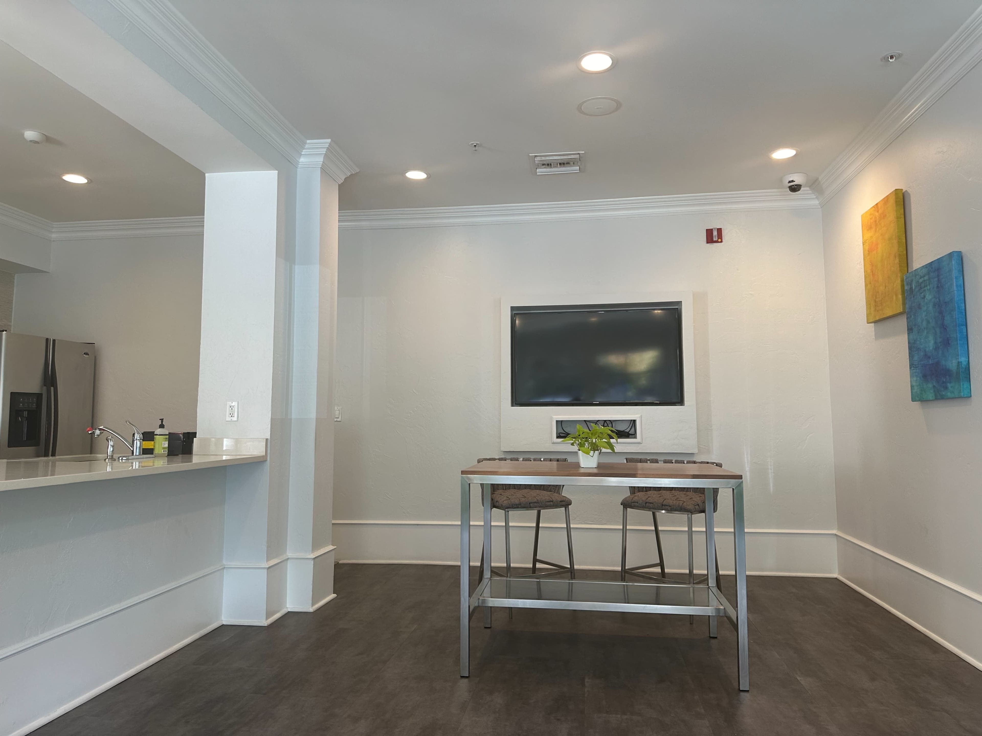 A modern kitchen area features a countertop, two bar stools, and a wall-mounted television in a bright, minimalist setting.