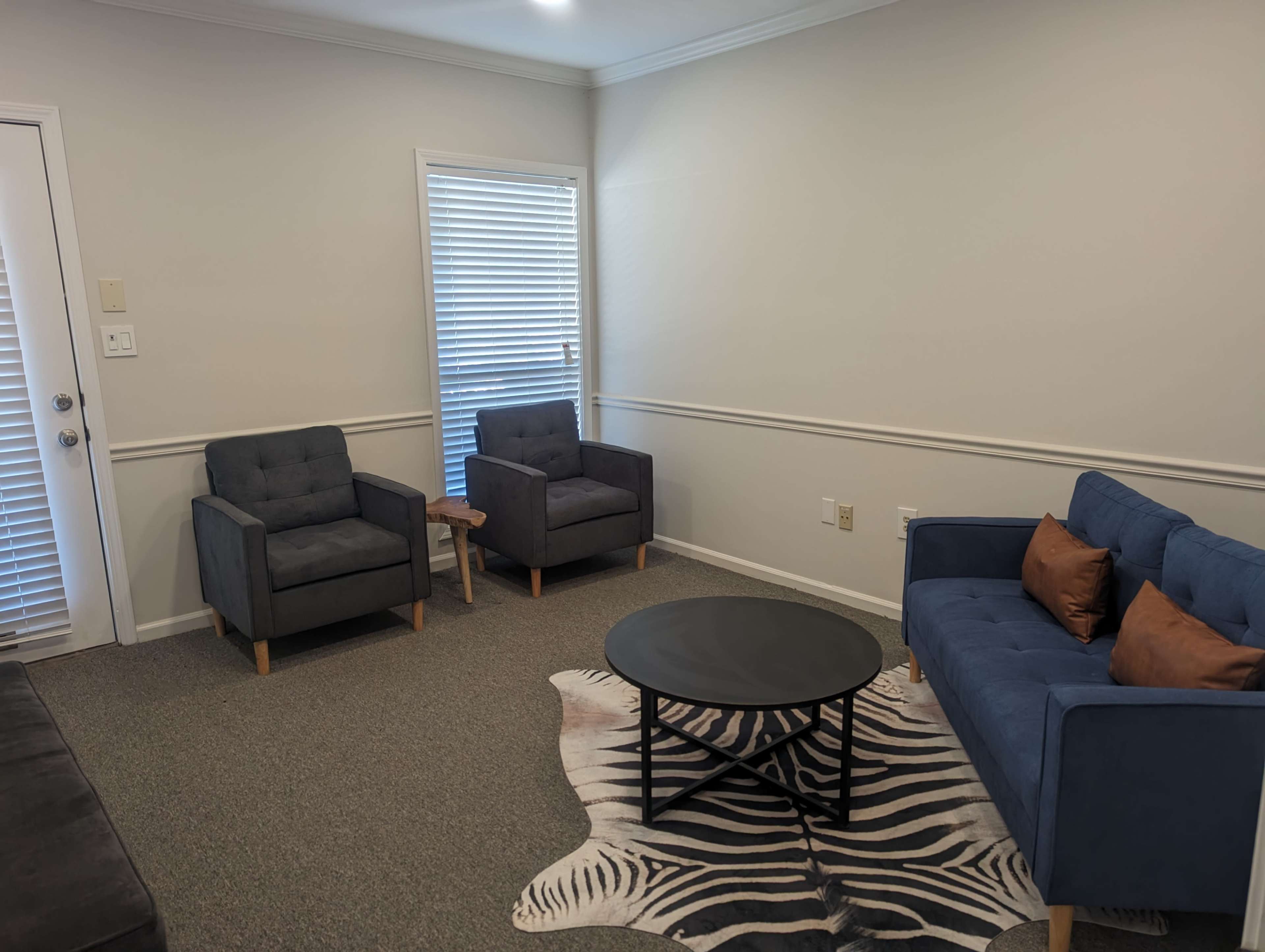 The image shows a living room with two gray armchairs, a blue sofa with decorative cushions, a round black coffee table, and a zebra-print rug.
