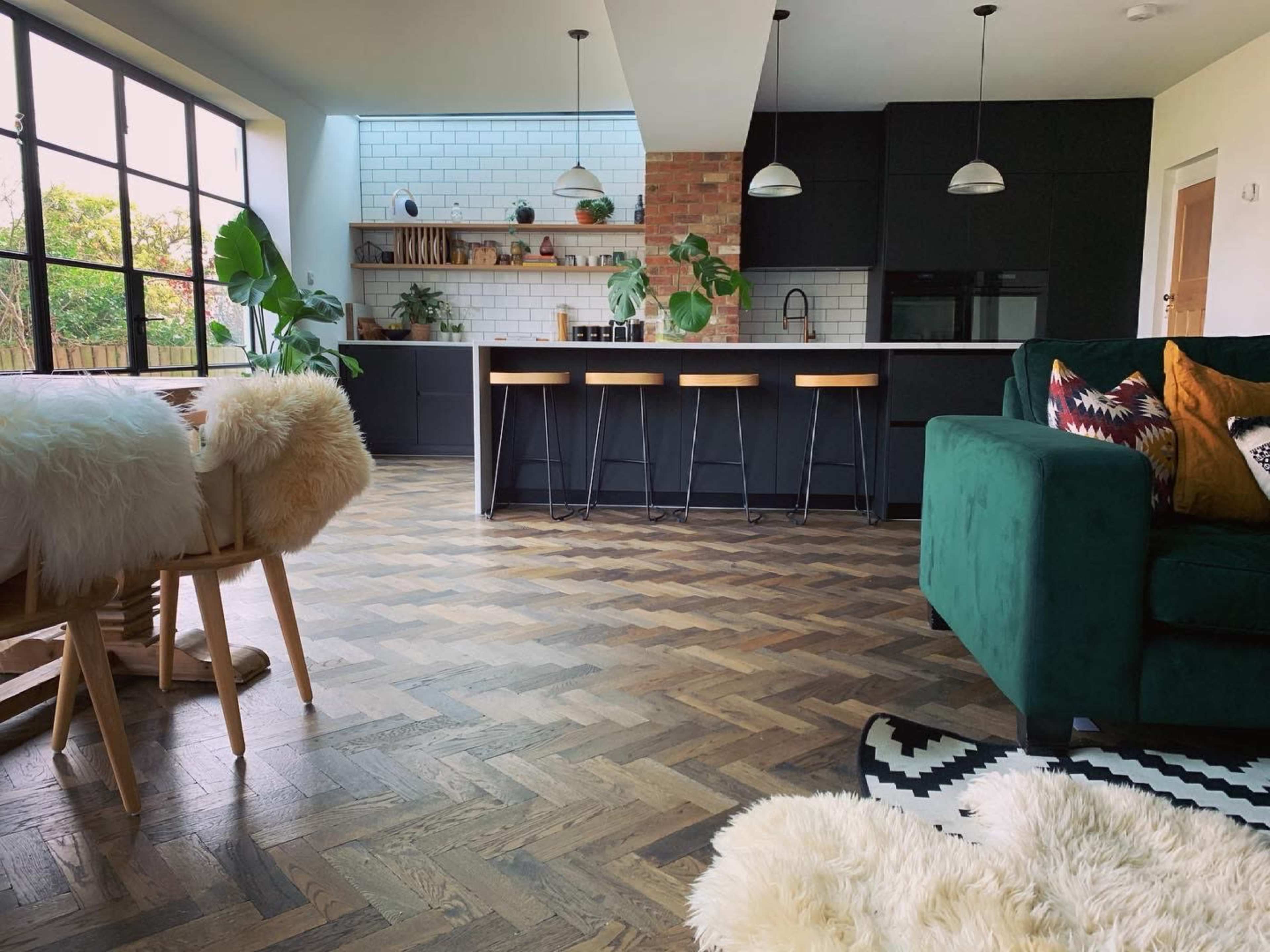 A modern kitchen and living area featuring a herringbone wood floor, black cabinetry, a large window allowing natural light, and a green sofa.