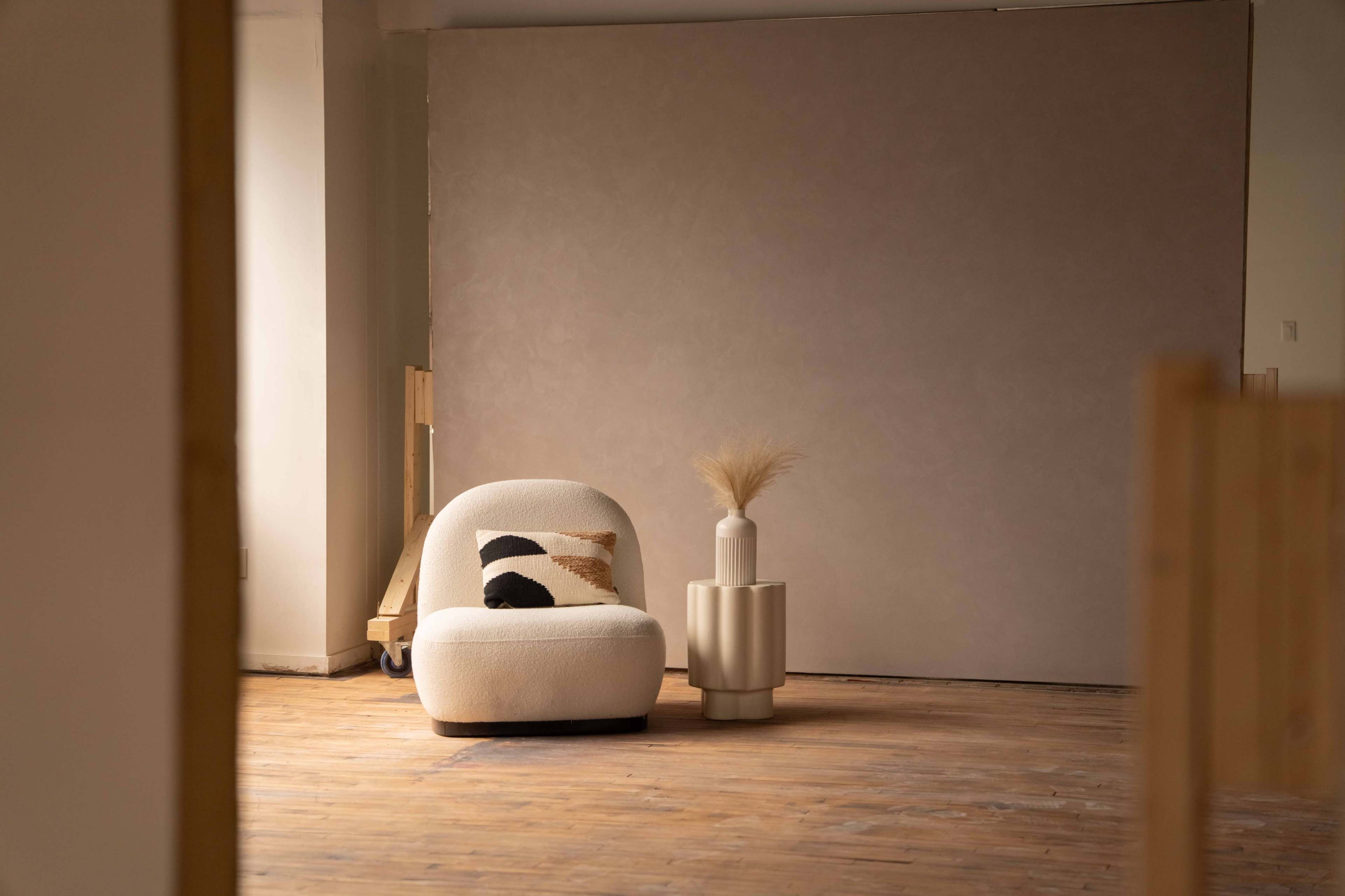 The image shows a cozy seating area with a curved chair and a round side table adorned with a decorative vase and dried plants, set against a neutral wall and wooden floor.