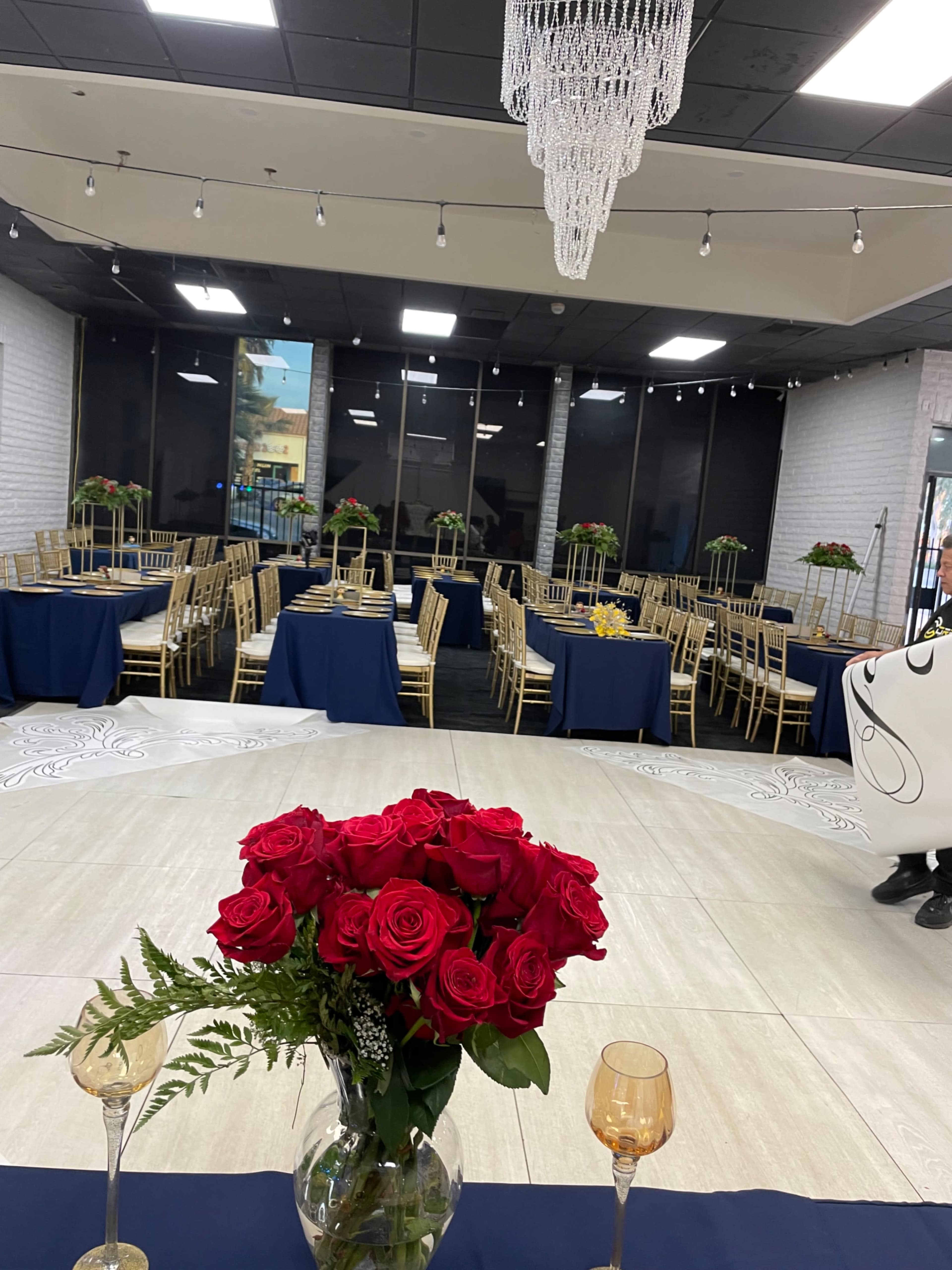 A banquet hall setup with tables covered in navy cloth, adorned with red roses, and gold chairs arranged around the space.